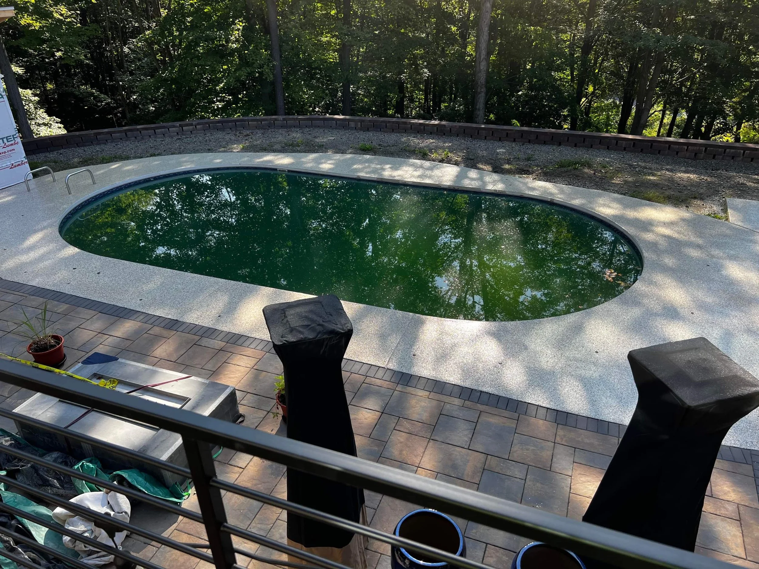 A small oval swimming pool with greenish water surrounded by a paved patio area, with two black covered outdoor heaters or equipment on the sides, in a backyard setting with trees in the background.
