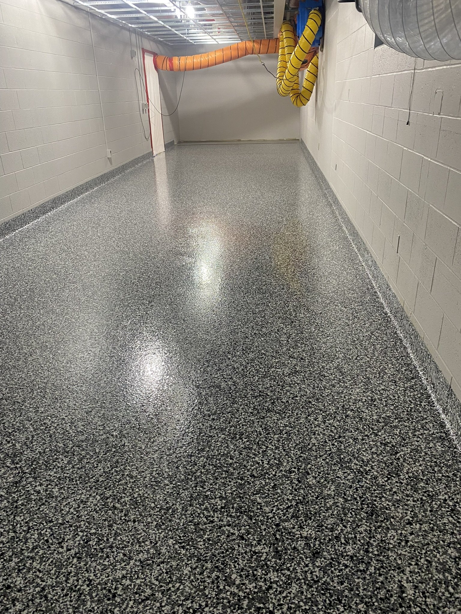 Empty industrial hallway with speckled epoxy flooring, exposed ceiling pipes, and ventilation ducts.