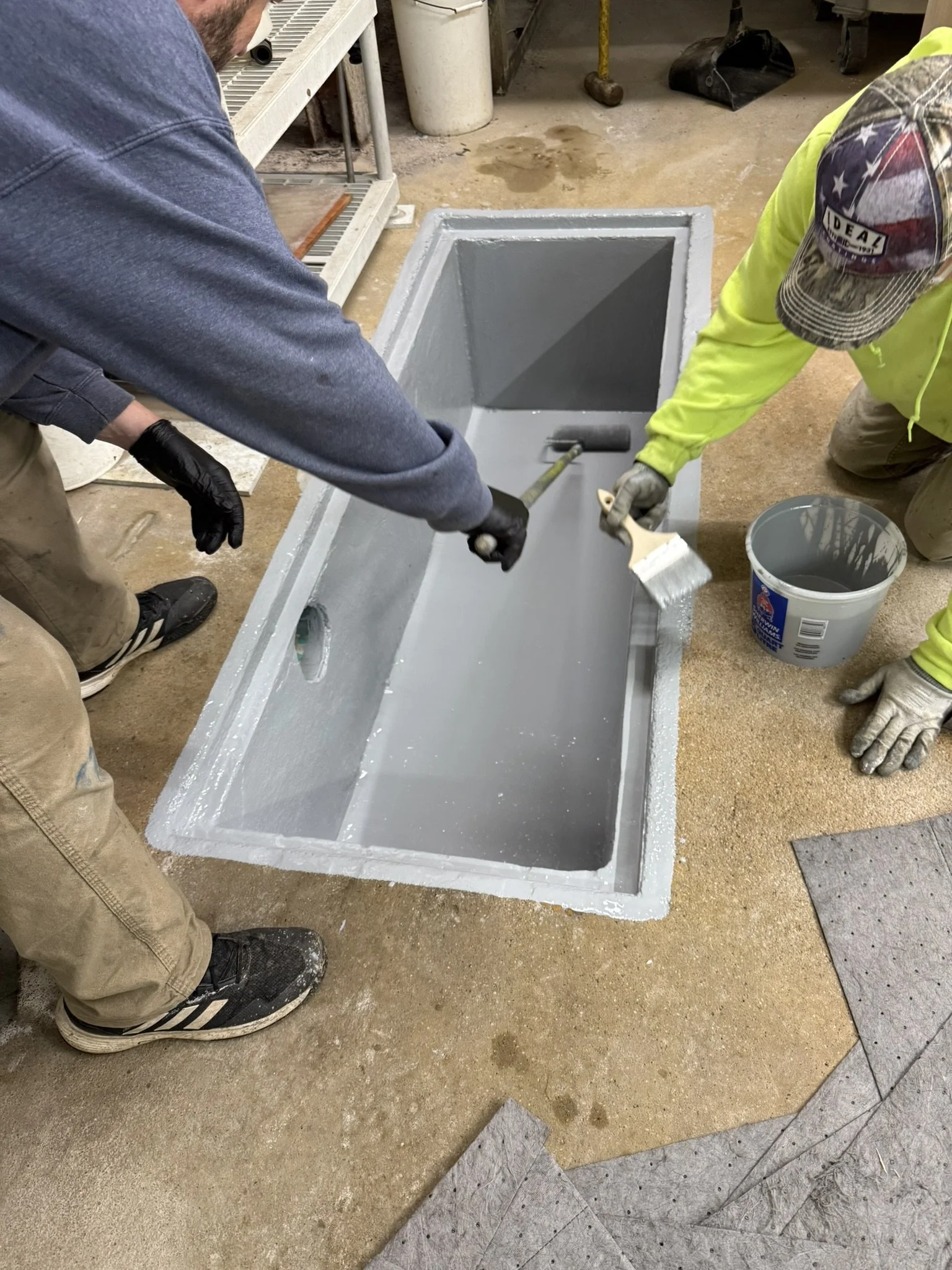 Two people applying gray paint to a concrete basin with brushes and a roller in a workshop setting.
