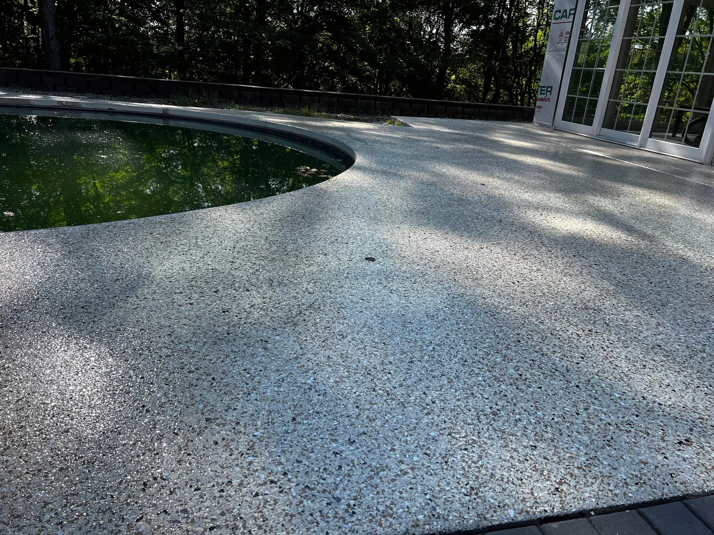 Concrete patio with speckled texture adjacent to a round swimming pool with murky water, surrounded by trees and a building with large windows.