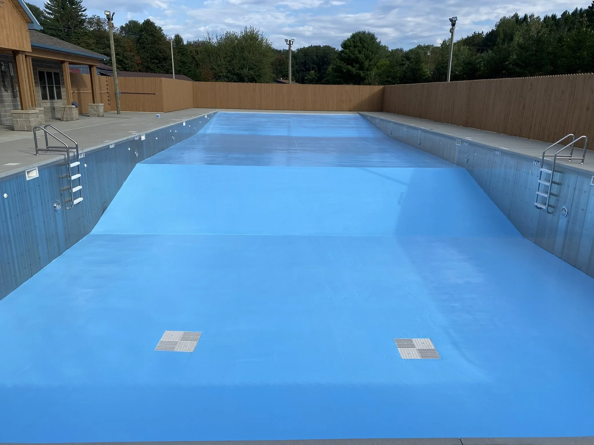 Empty outdoor swimming pool with light blue painted interior, surrounded by concrete decking and wooden fence, with metal ladders on the sides.