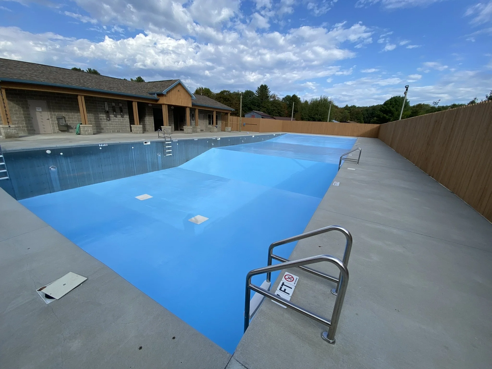 Empty outdoor swimming pool covered with blue covers, surrounded by concrete deck, with nearby building and cloudy sky.