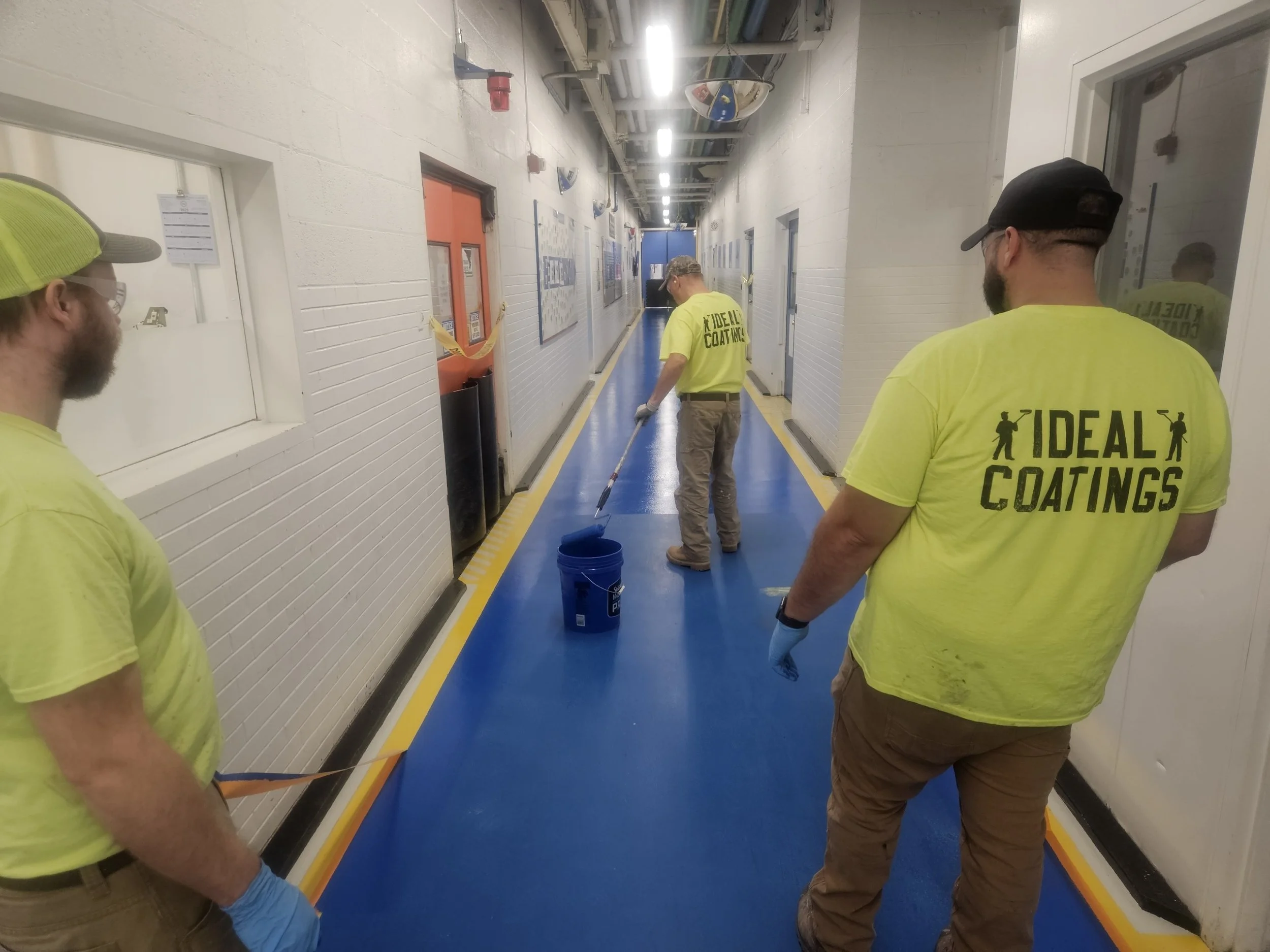Three men wearing neon yellow shirts with "Ideal Coatings" text, working in a hallway applying blue floor coating with rollers.