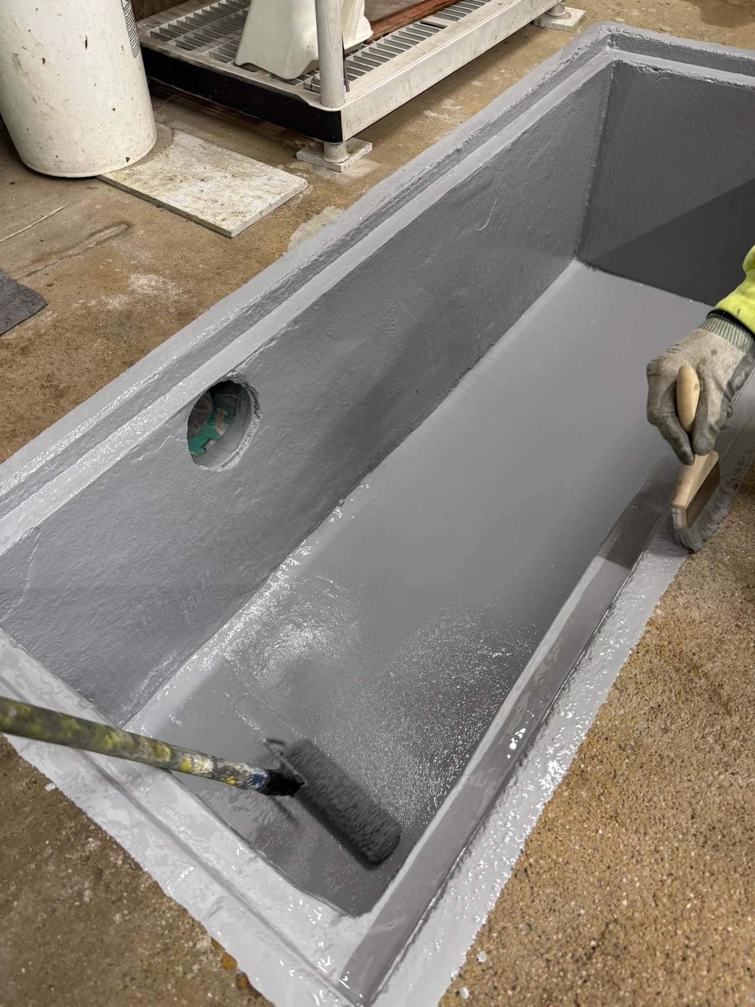 A person using a paint roller to apply gray coating to a rectangular concrete structure with an open top, possibly a drainage box or trough, inside a workshop area.
