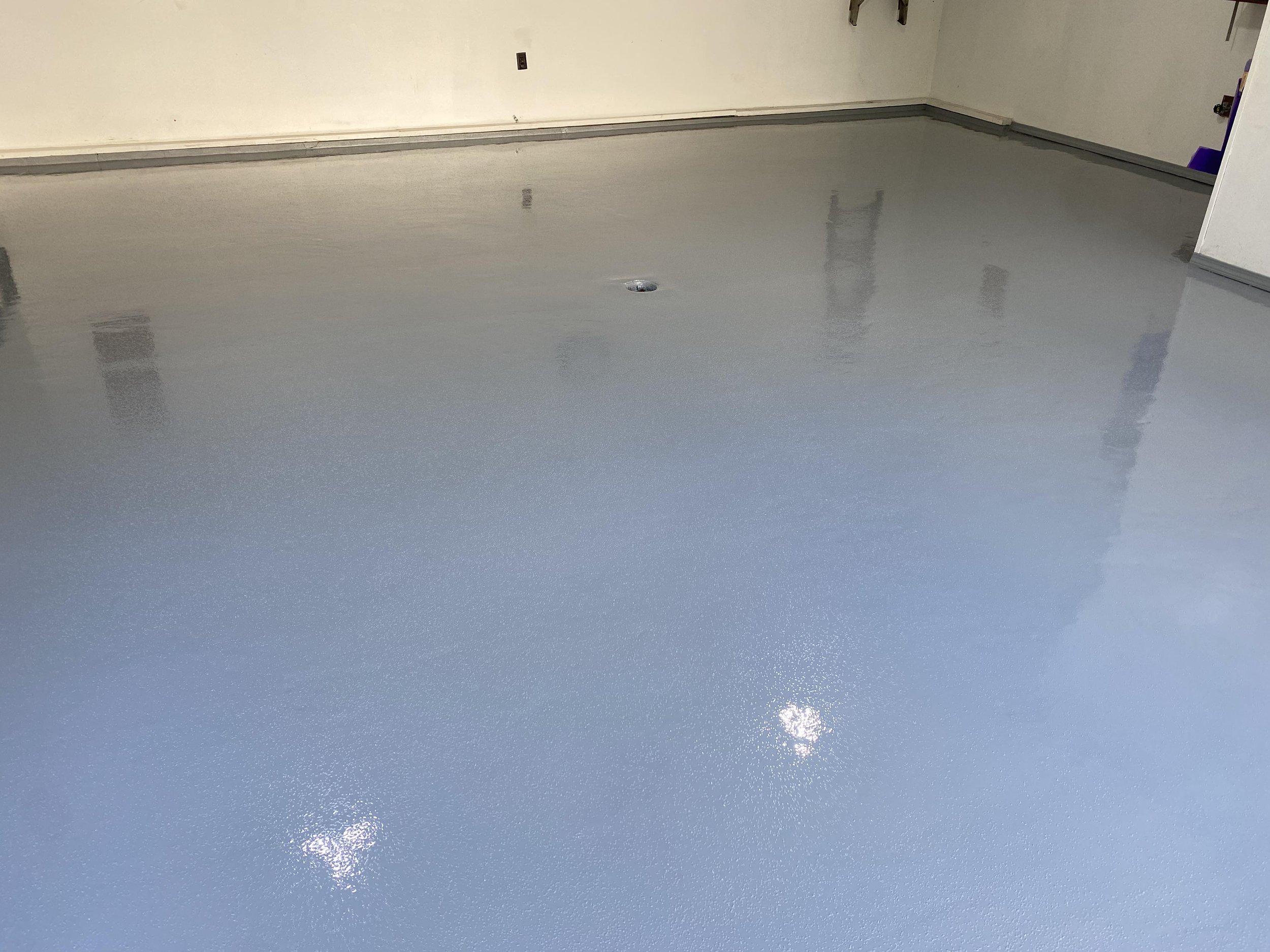 Freshly coated garage floor with a gray epoxy finish, reflecting light; bare white walls in the background.