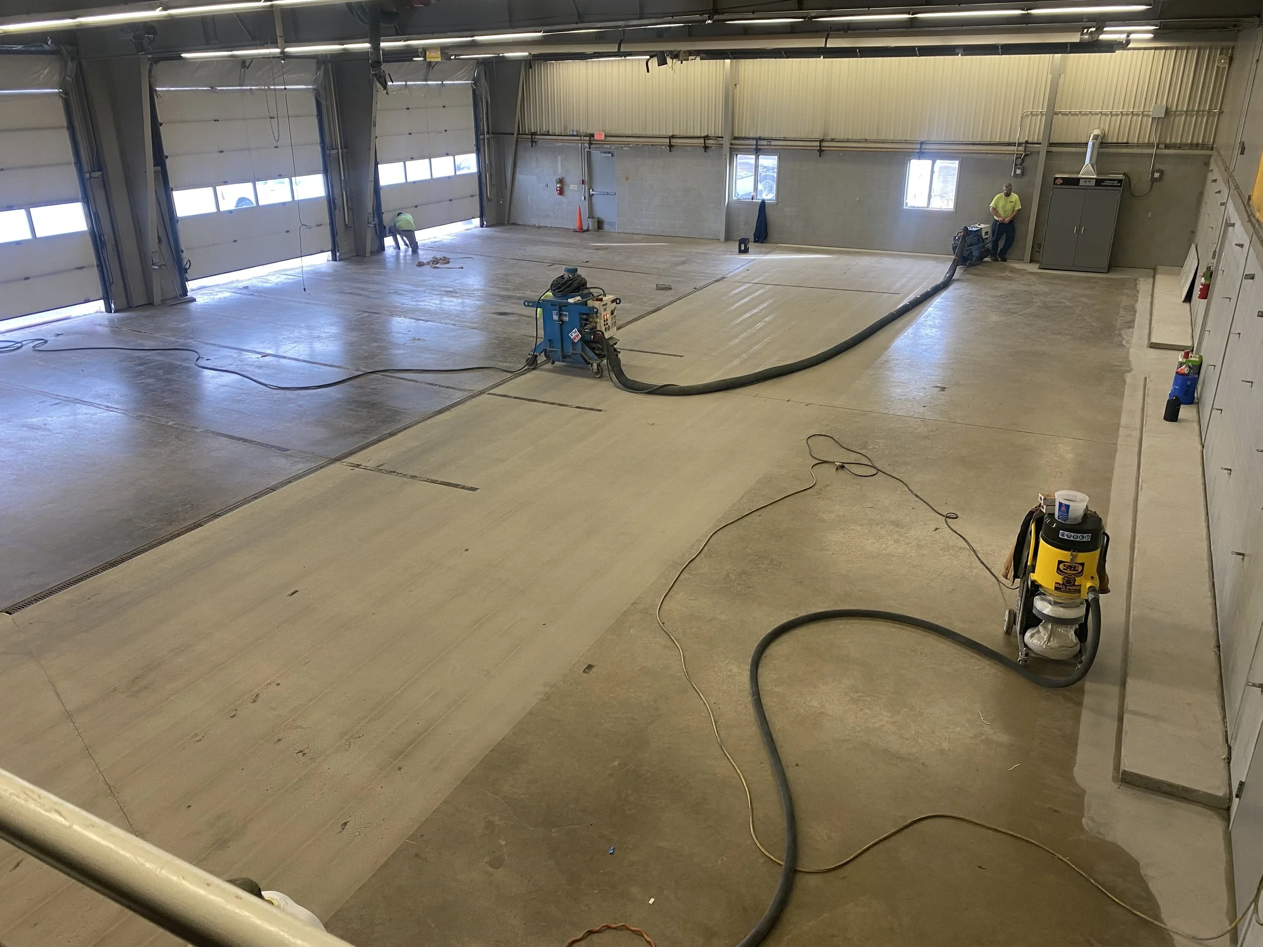 Large industrial garage with concrete floor being cleaned or prepared by workers using equipment.