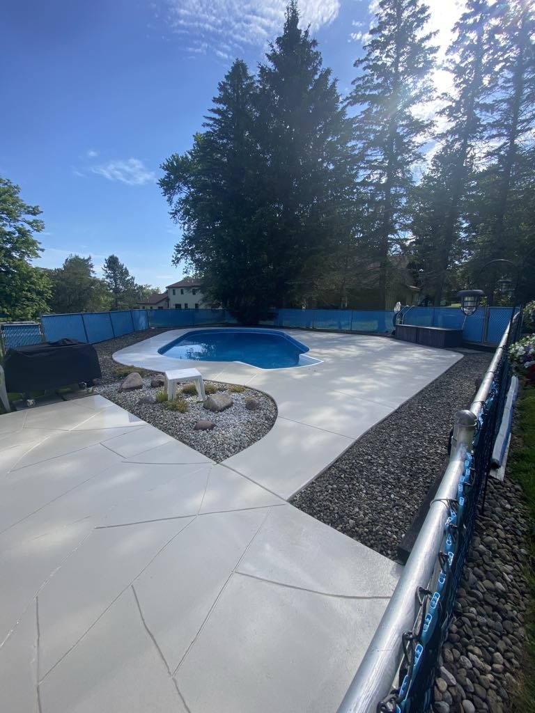 Backyard swimming pool with cement walkway, surrounded by trees and a blue fence.