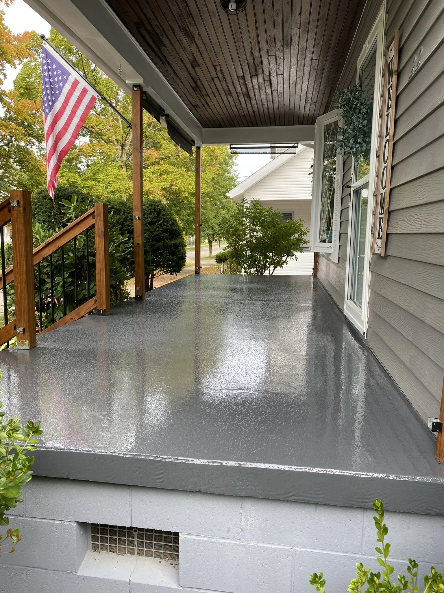 A porch with a glossy gray surface, wood railing, and a "Welcome" sign. An American flag is displayed on a pole. The house has gray siding, a wooden ceiling, and an open window. Greenery surrounds the area.