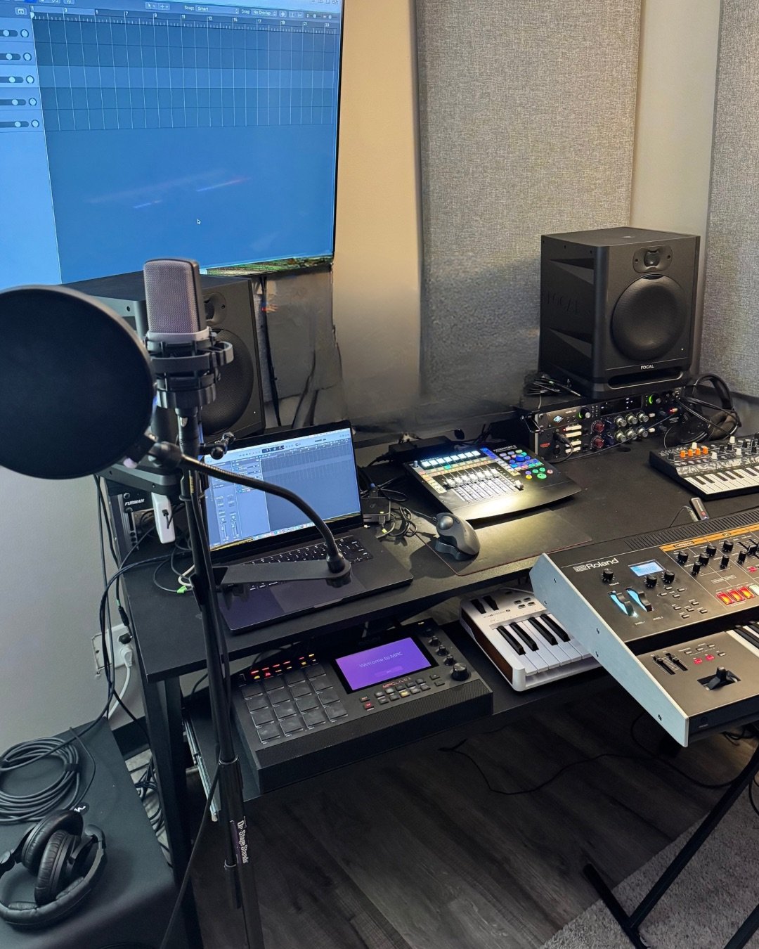 Music production studio with a computer, MIDI controllers, studio monitors, audio interface, microphone, and headphones on a desk.