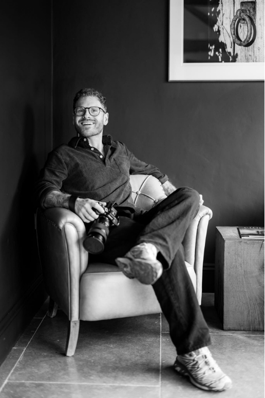 A man with glasses sitting on an armchair holding a camera in his right hand, smiling, in a room with dark walls, a framed picture on the wall, and a small table beside the chair.