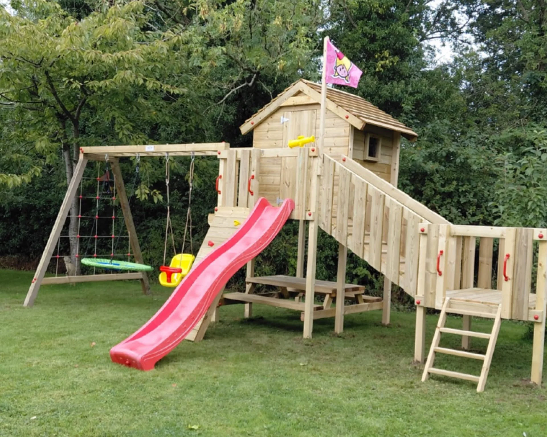 Wooden playhouse with two slides, one red and one green, in a backyard with grass and a fence.