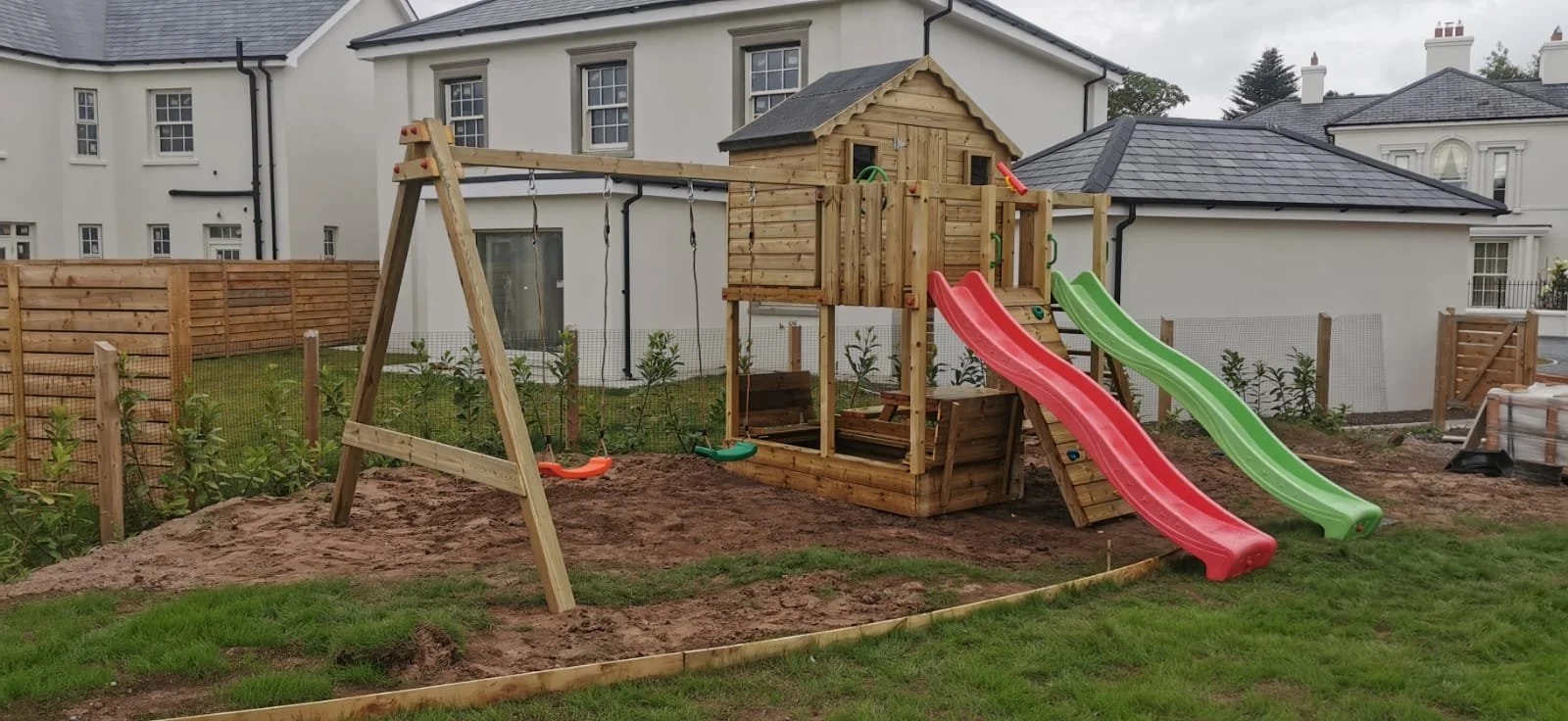 Children's outdoor playset with swings, dual slides (red and green), and a wooden playhouse in a backyard.