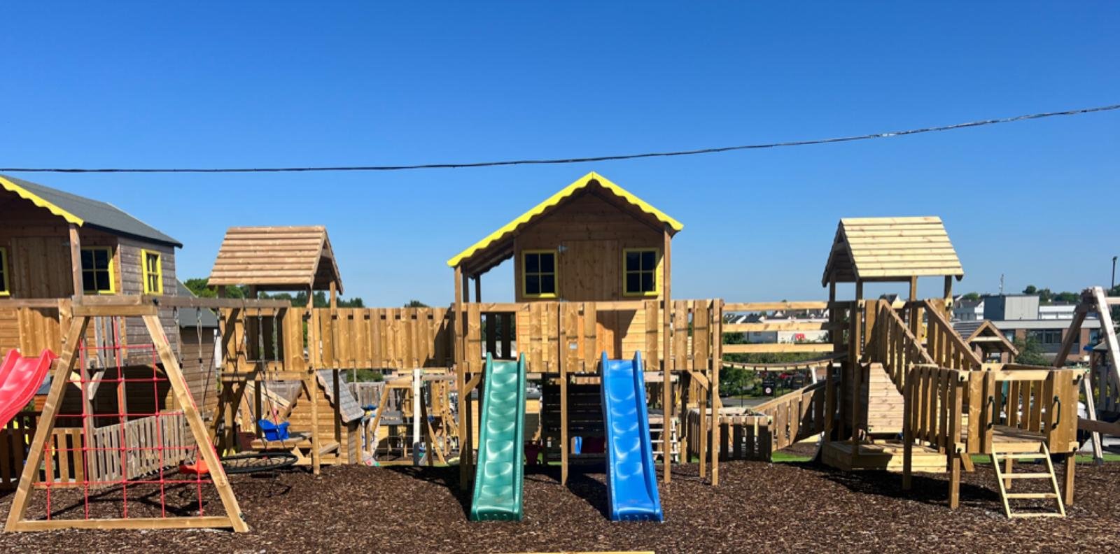 A backyard playground with wooden play structures, slides, swings, and a climbing net under a clear blue sky.