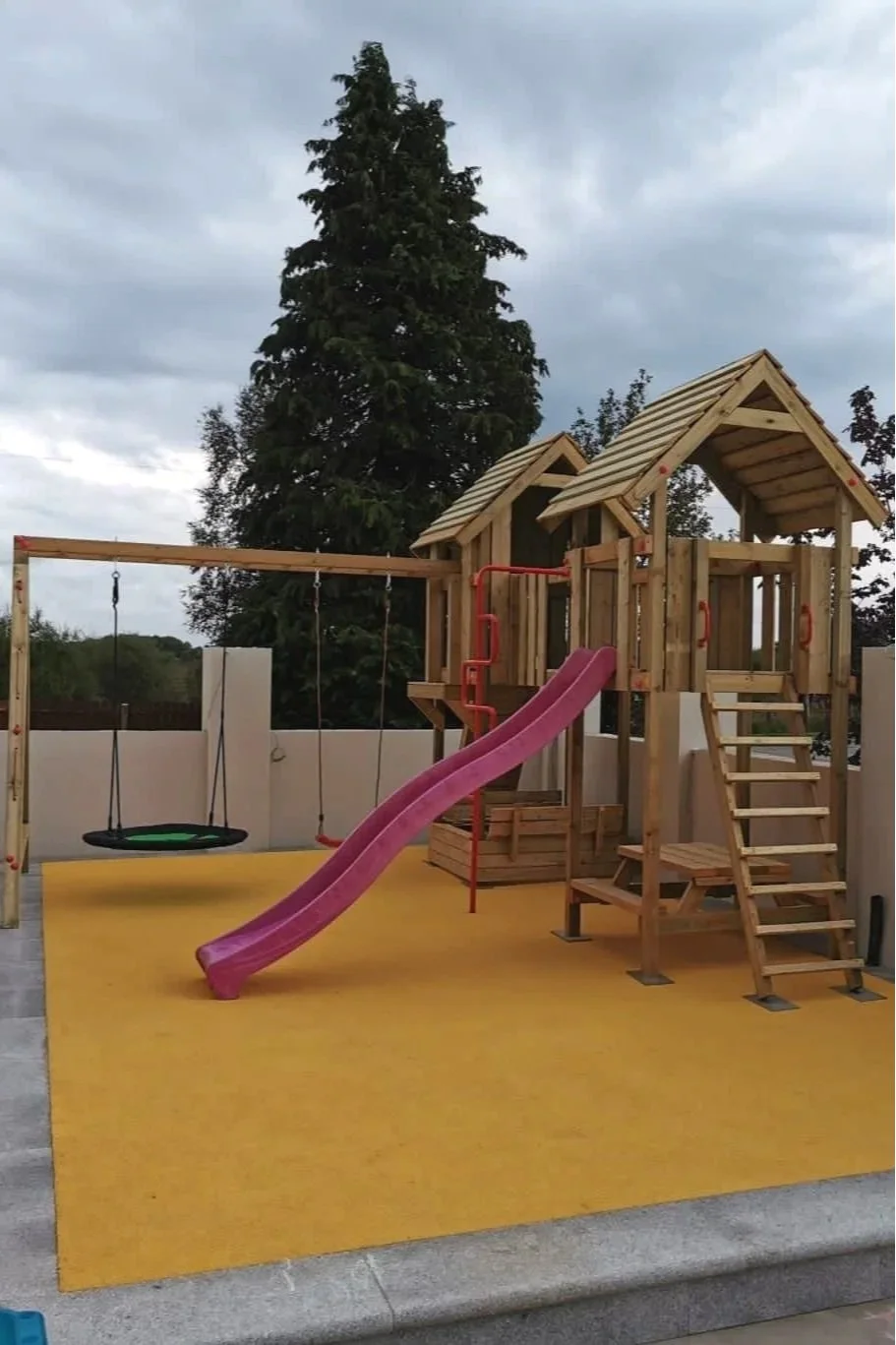 A children's playground with a wooden play house, pink slide, swings, and climbing ladder on a yellow soft surface, with trees and cloudy sky in the background.