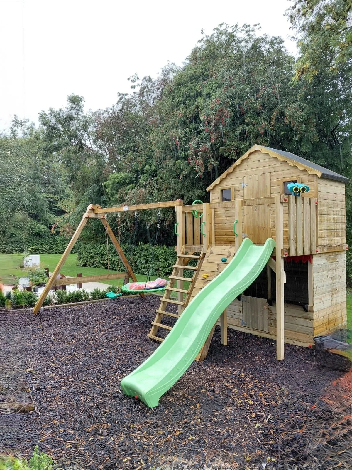 A wooden playground set with slides and swings, situated in a park with trees and a gravel surface.