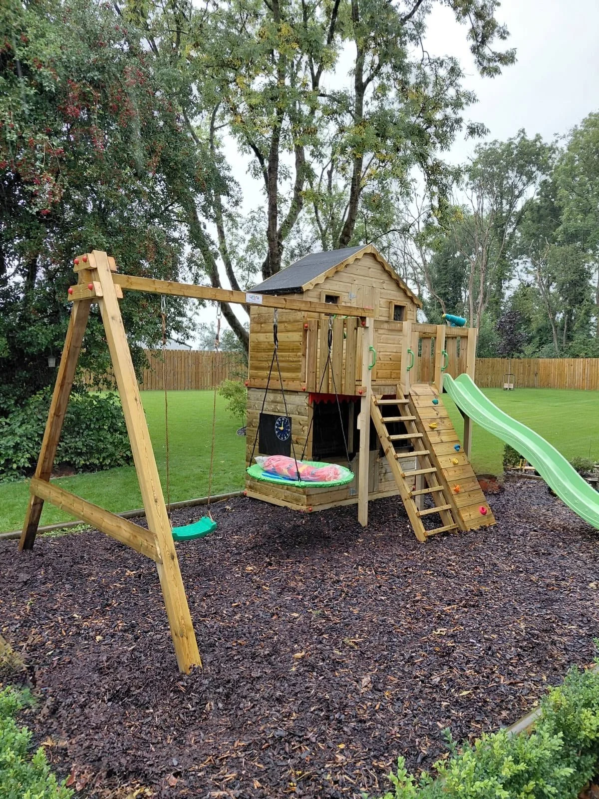 A wooden children's playset with a slide, climbing wall, swings, and a small covered platform in a playground surrounded by trees and bushes.