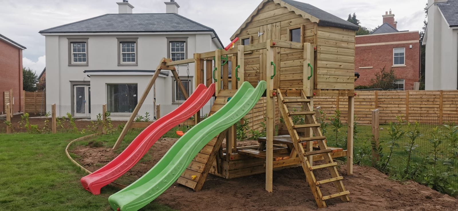 Wooden playhouse with two slides, one red and one green, in a backyard with grass and a fence.
