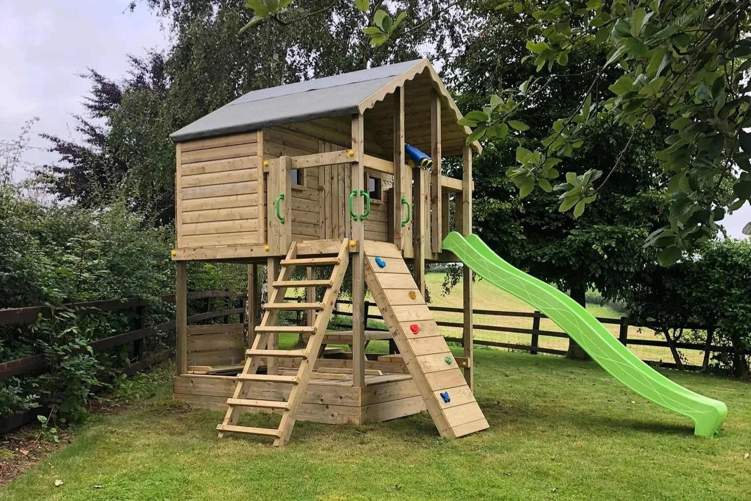 Wooden outdoor playhouse with a green slide and climbing wall, situated on a grassy yard with trees and a wooden fence in the background.