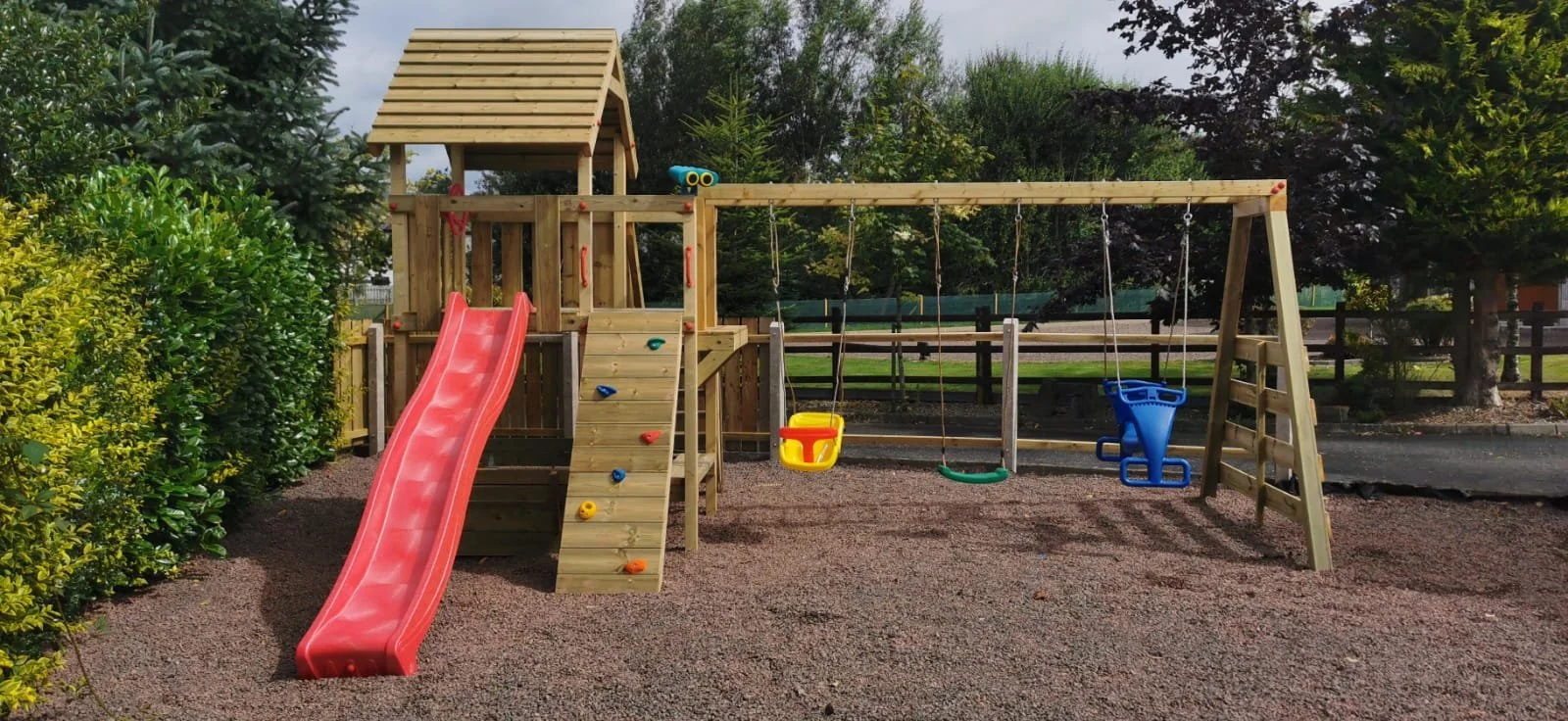 A wooden playground set with slides and swings, situated in a park with trees and a gravel surface.