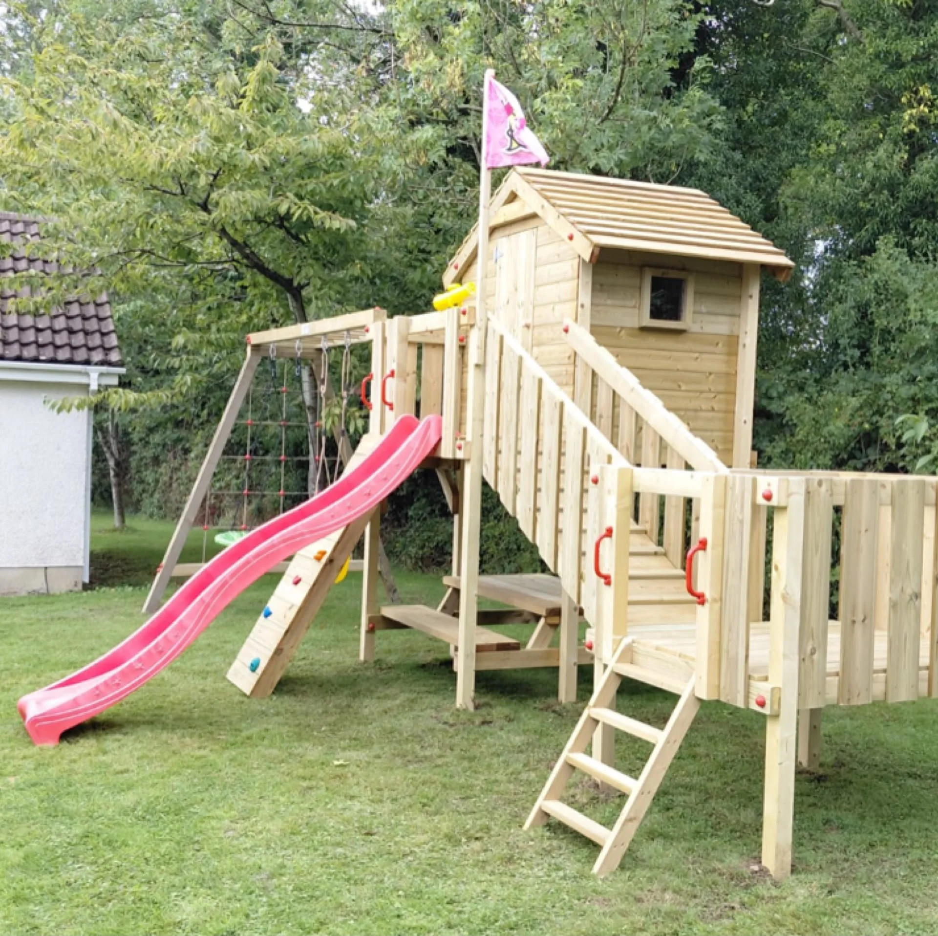 Children's outdoor playset with swings, dual slides (red and green), and a wooden playhouse in a backyard.