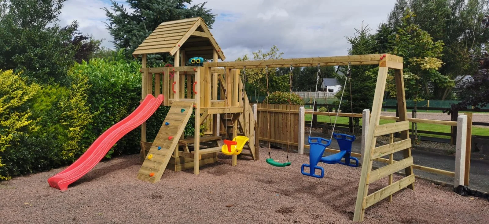 A wooden children's playset with a slide, climbing wall, swings, and a small covered platform in a playground surrounded by trees and bushes.