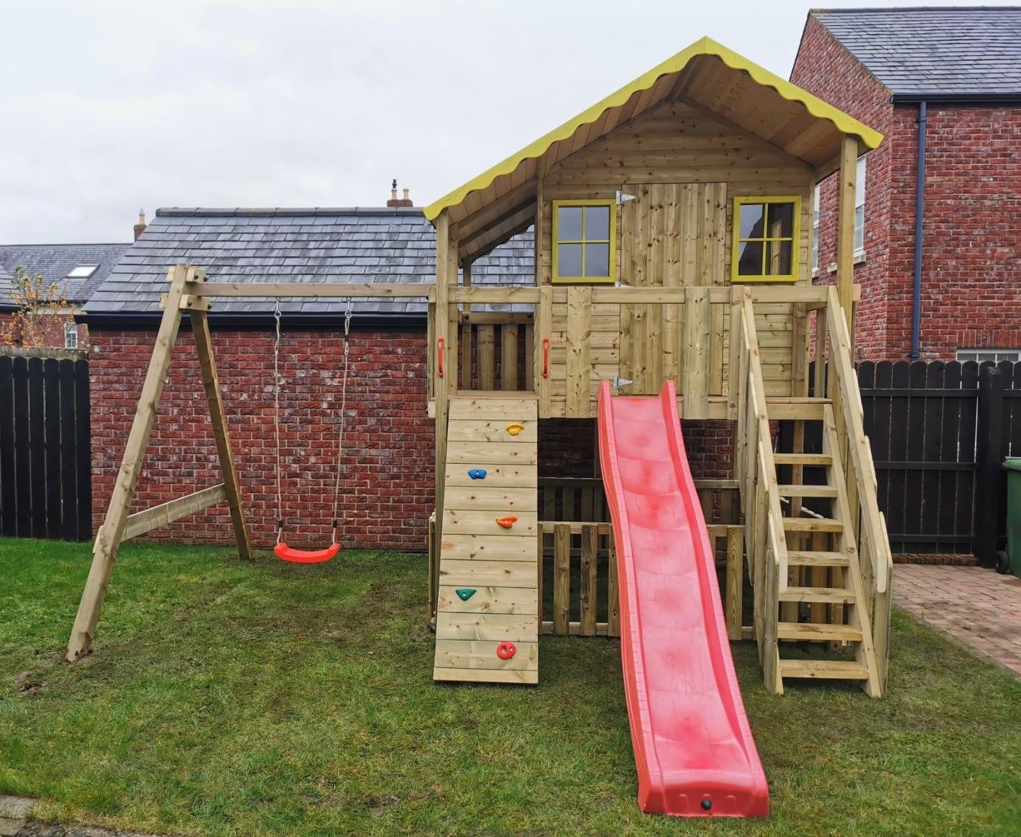 A wooden playhouse with a slide, climbing wall, and swingset in a backyard