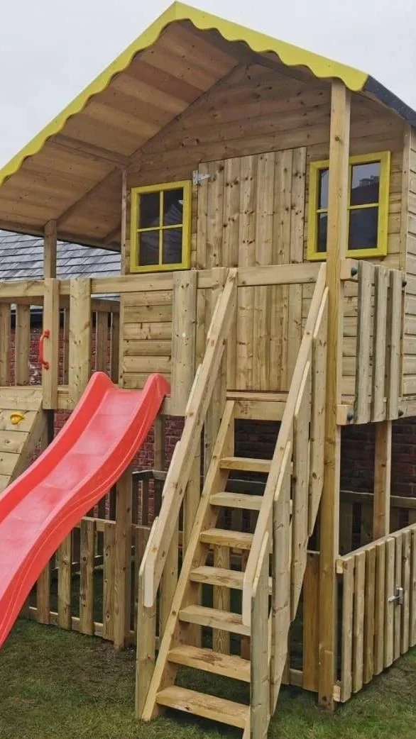 A wooden playhouse on stilts with a yellow roof trim, a red slide, and a wooden staircase with handrails. The playhouse has two small windows with yellow trim, and a small fence surrounds the structure.