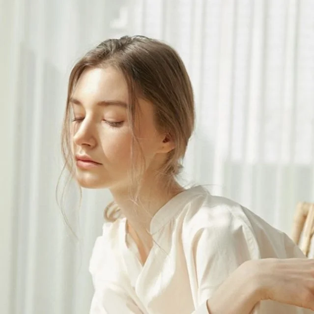 A young woman with closed eyes and relaxed expression, sitting in front of a sheer curtain in natural light, wearing a light-colored top.