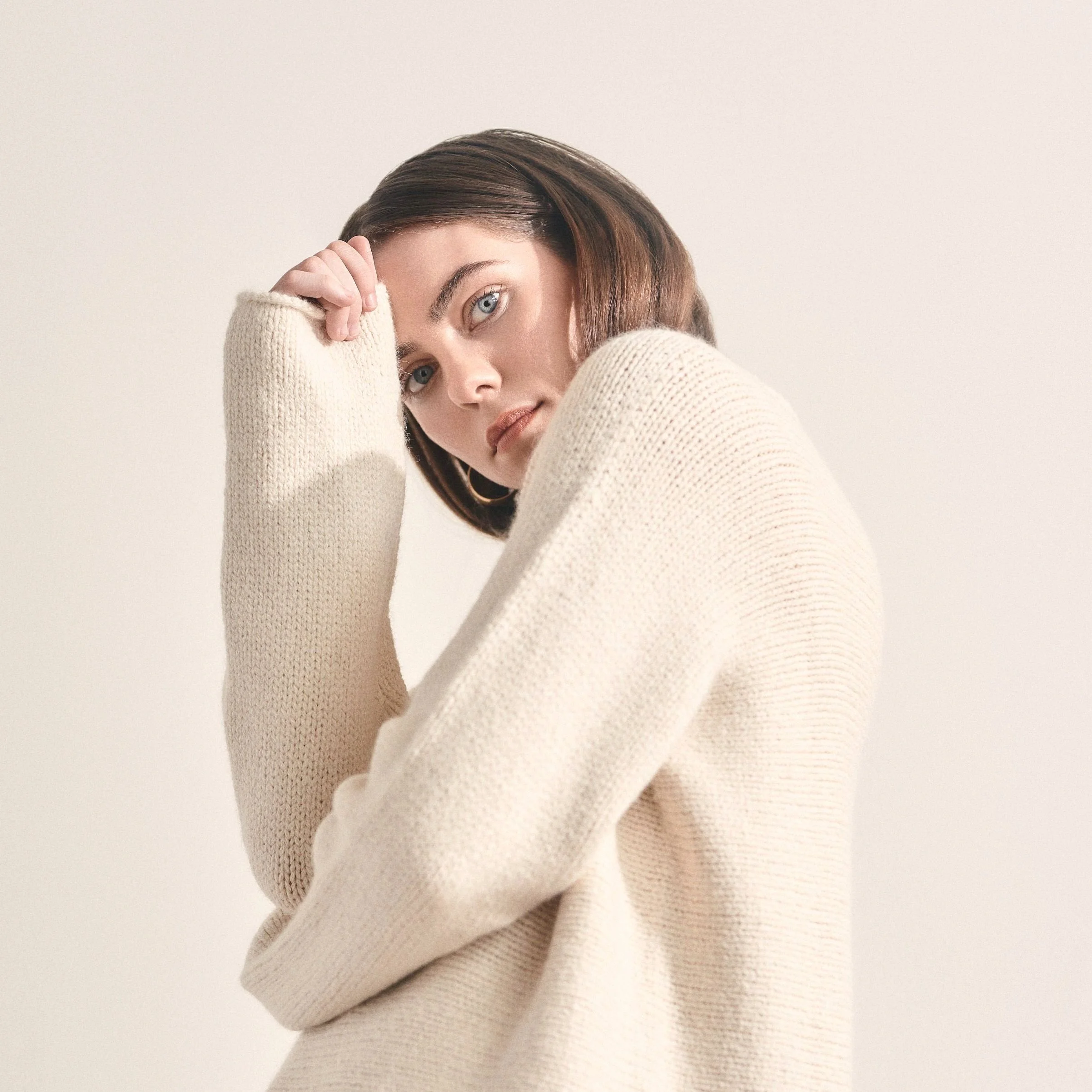 A young woman with brown hair and blue eyes wearing a cream-colored knit sweater, looking directly at the camera with her hand resting on her forehead.