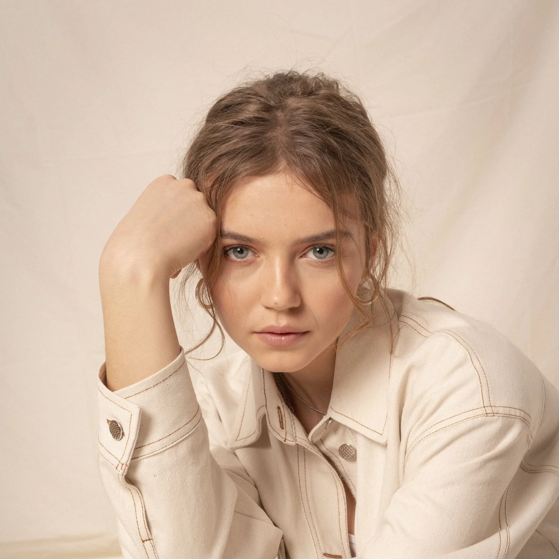A young woman with curly, shoulder-length brown hair and blue eyes leaning her head on her left arm, wearing a cream-colored jacket against a neutral background.