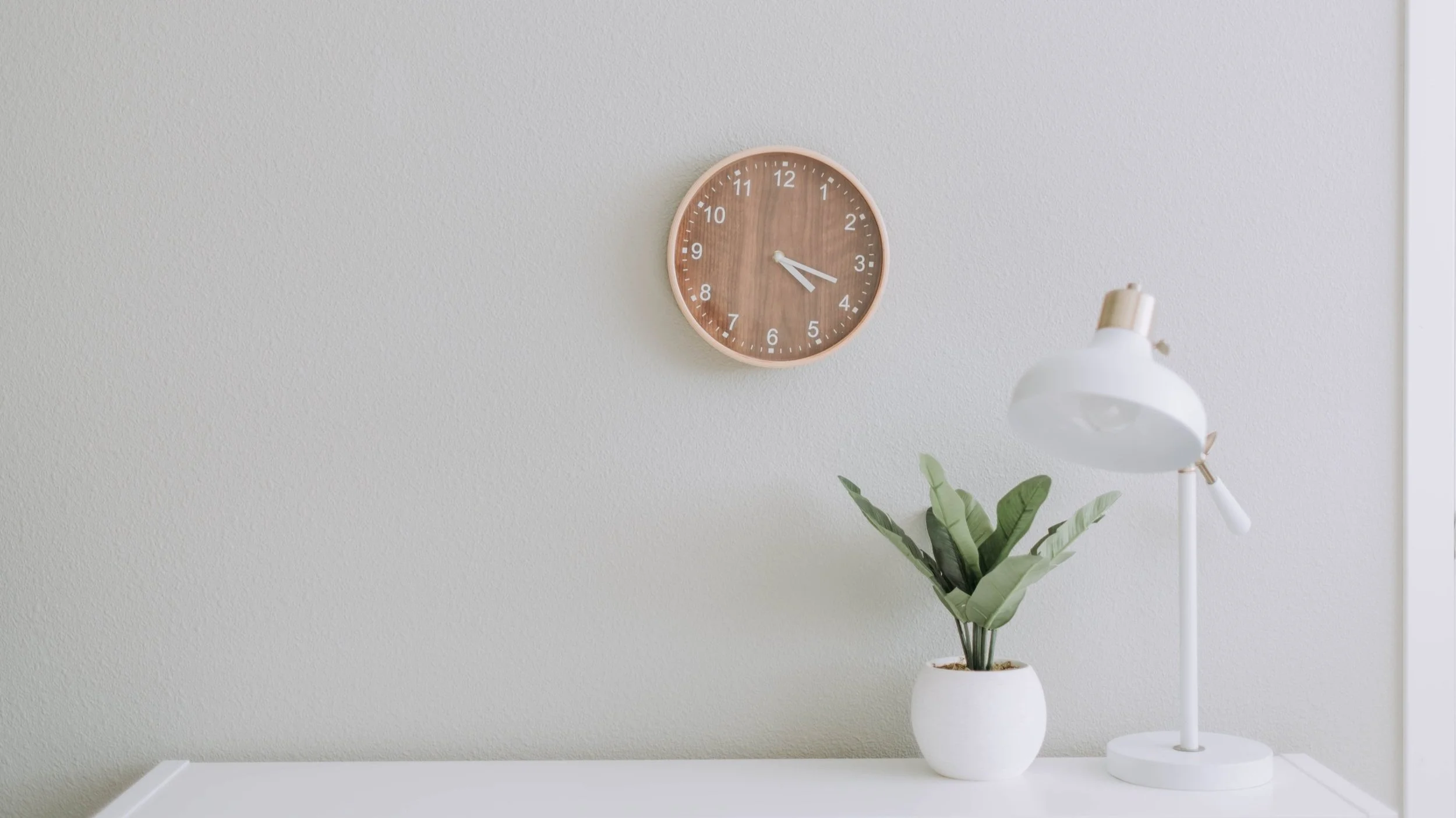 A white desk with a white table lamp and green potted plant. A round wall clock with a wooden face hangs on a light gray wall.