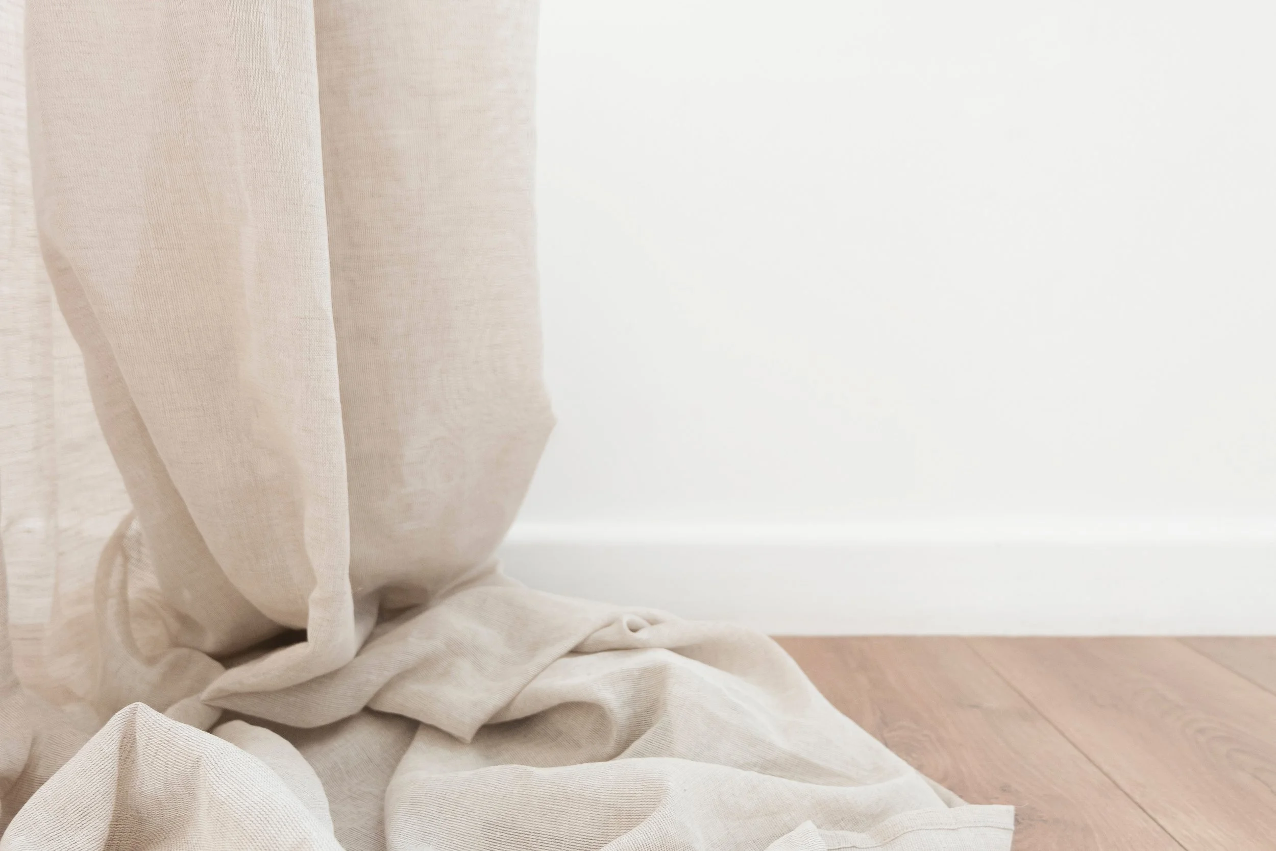 Close-up of beige, semi-transparent curtains on wooden floor near white wall.
