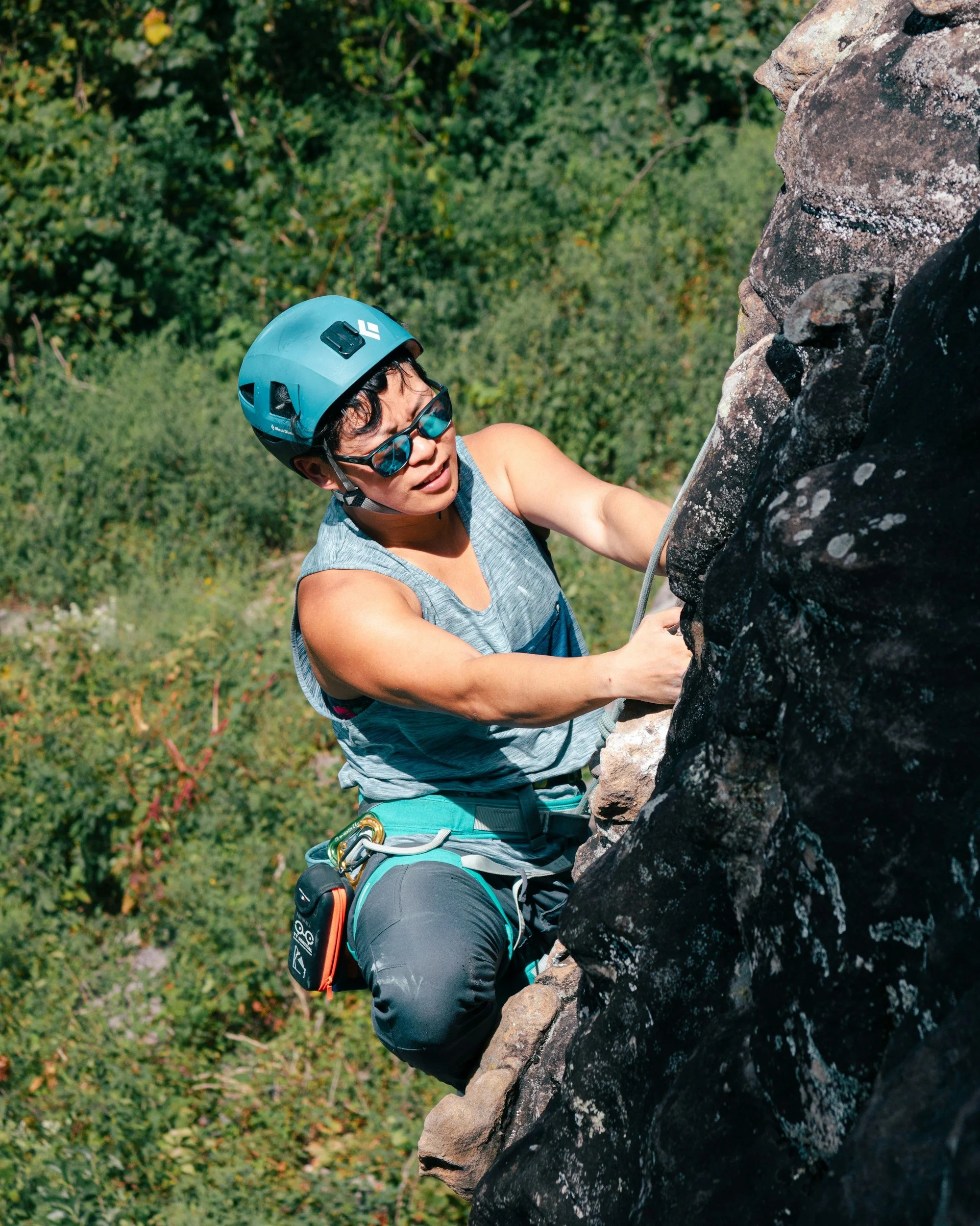 Person wearing a red climbing helmet, holding various climbing cams and carabiners, sitting on rocks with a rock wall in the background.
