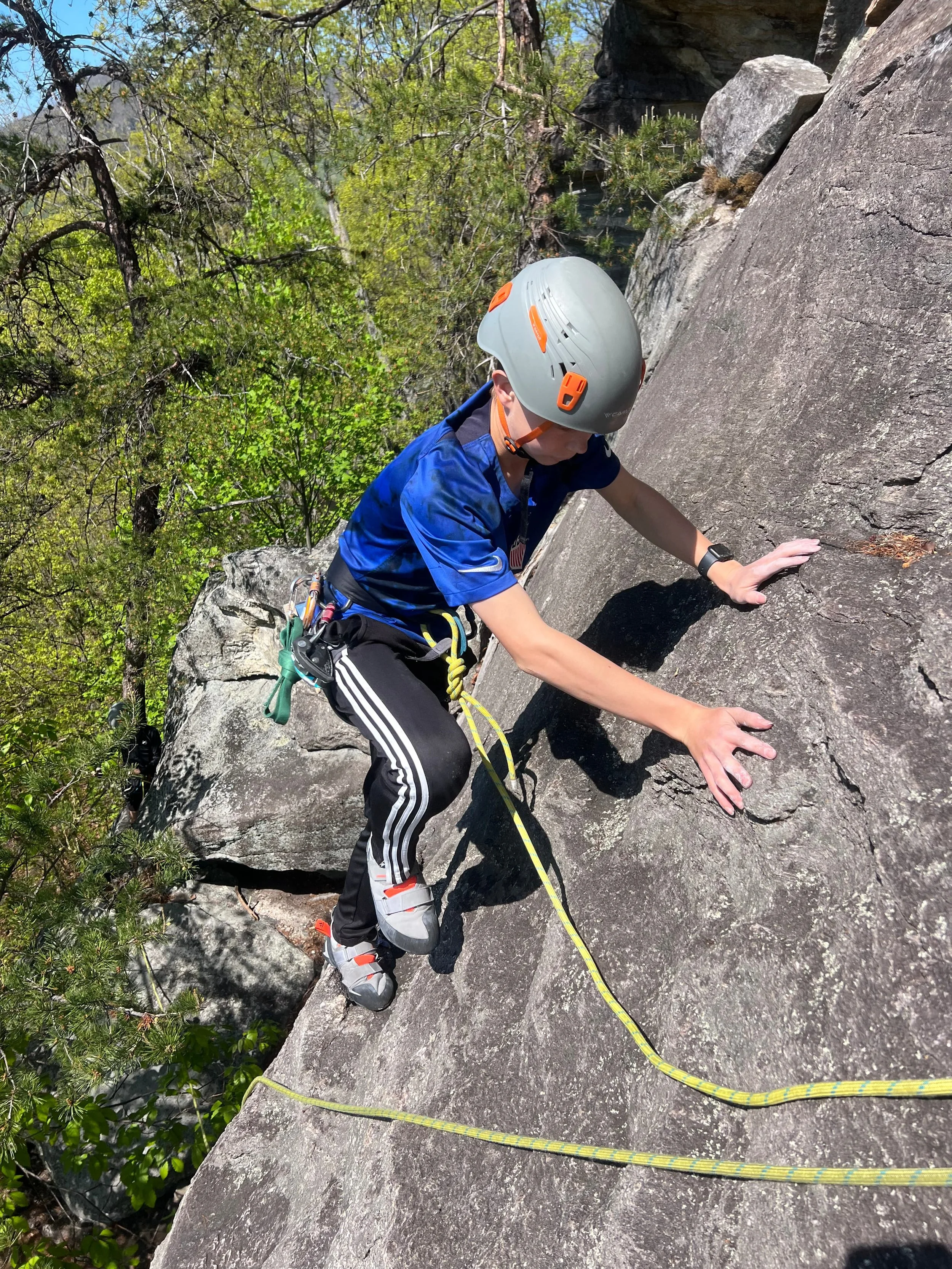 A person wearing climbing gear, a helmet, and sports clothes is rock climbing outdoors on a large rock surface, with trees and blue sky in the background.