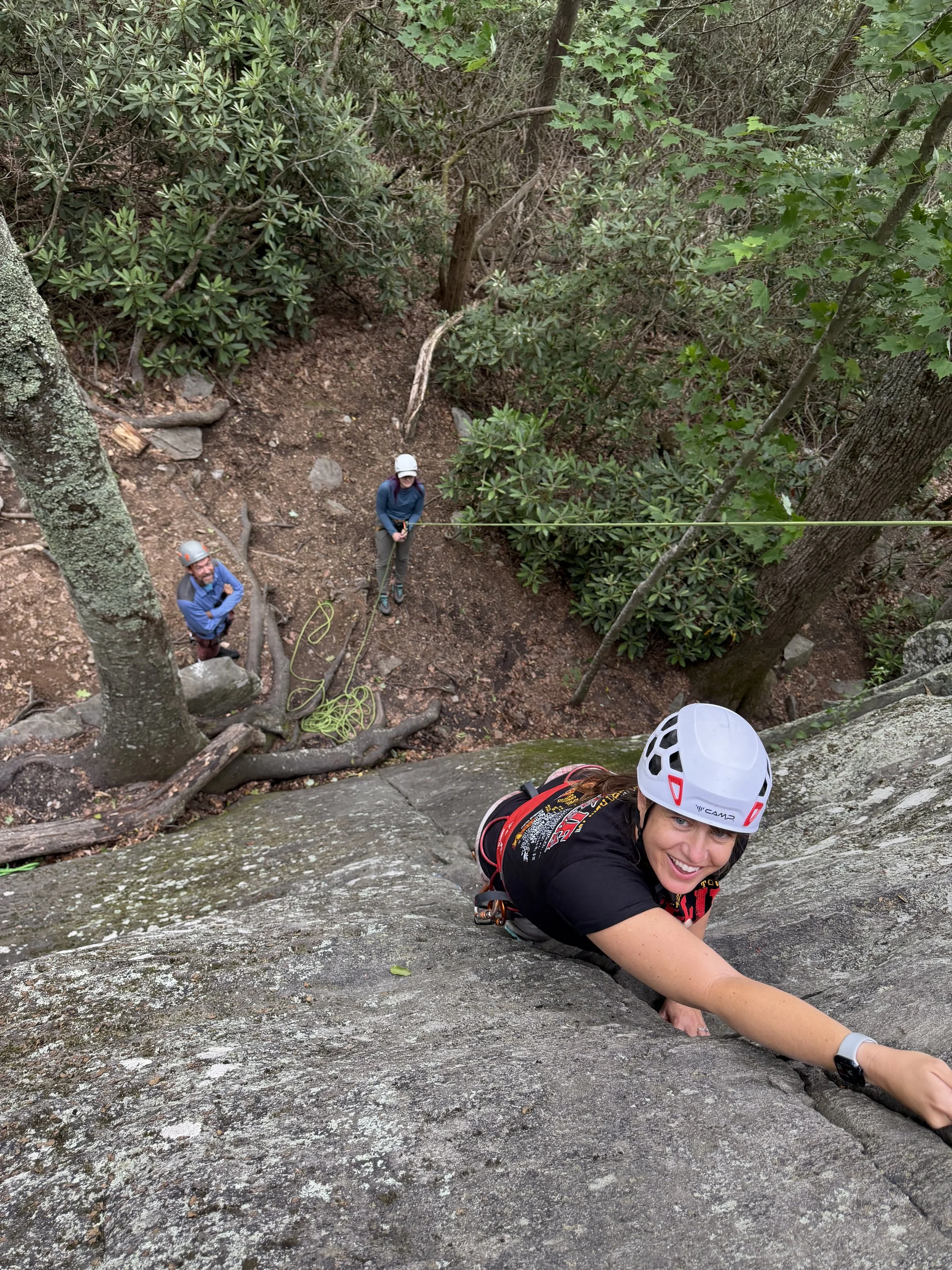 Young woman with helmet and climbing gear ascending a rock face with a smile, viewed from above, with two people and a forested area below.