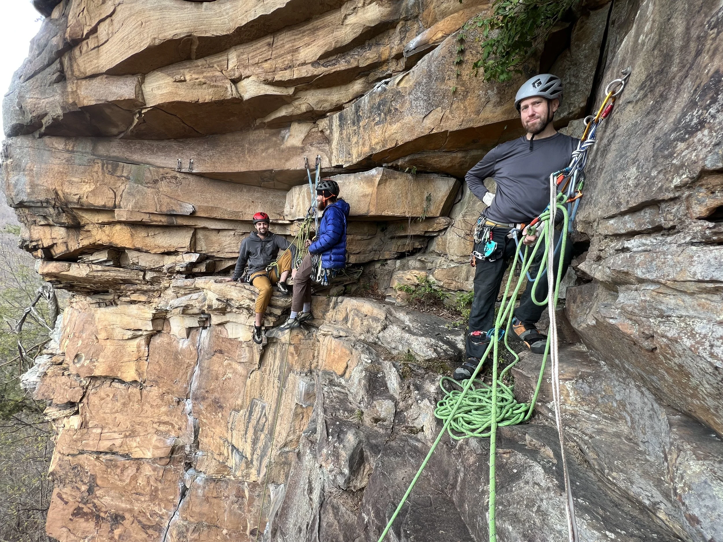 Three rock climbers on a ledge, wearing harnesses and helmets, with ropes and climbing gear, on a rocky cliff face.