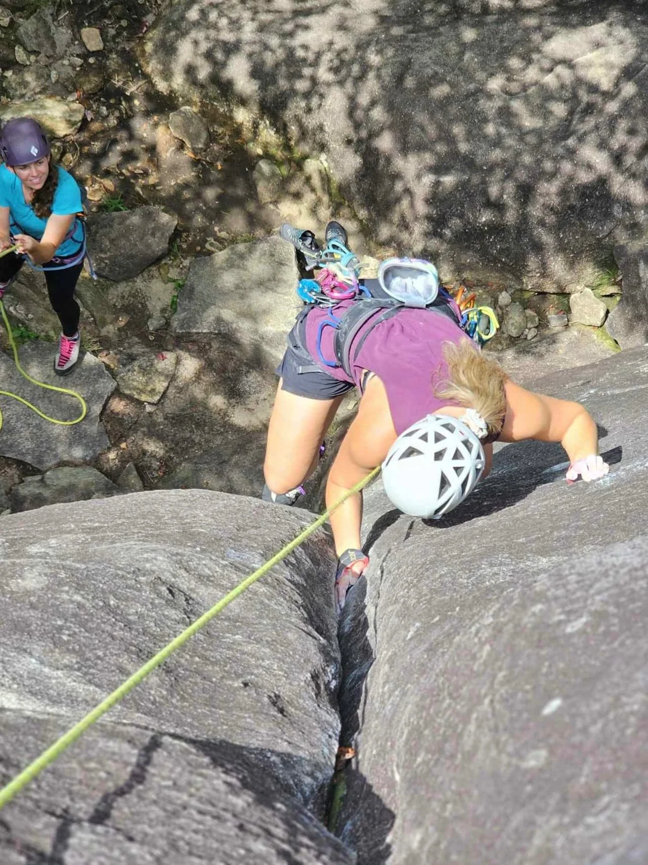 A woman rock climbing on a crack in a large rock face, wearing a helmet, harness, and climbing gear, with an assistant belaying her, outdoors in a rocky area.