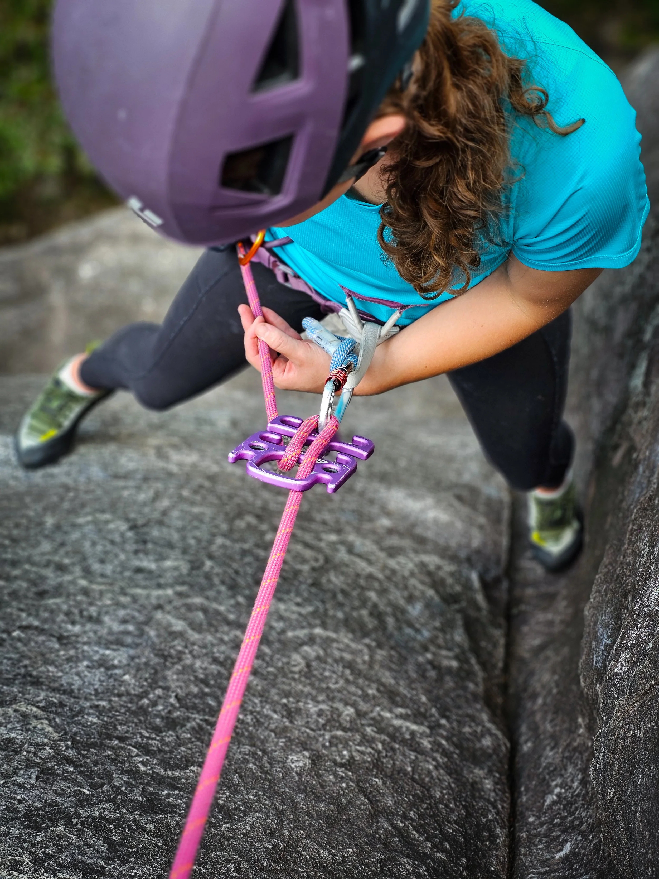 Rock climber using a belay device with a purple helmet and turquoise shirt, focusing on securing the rope on a rocky surface.