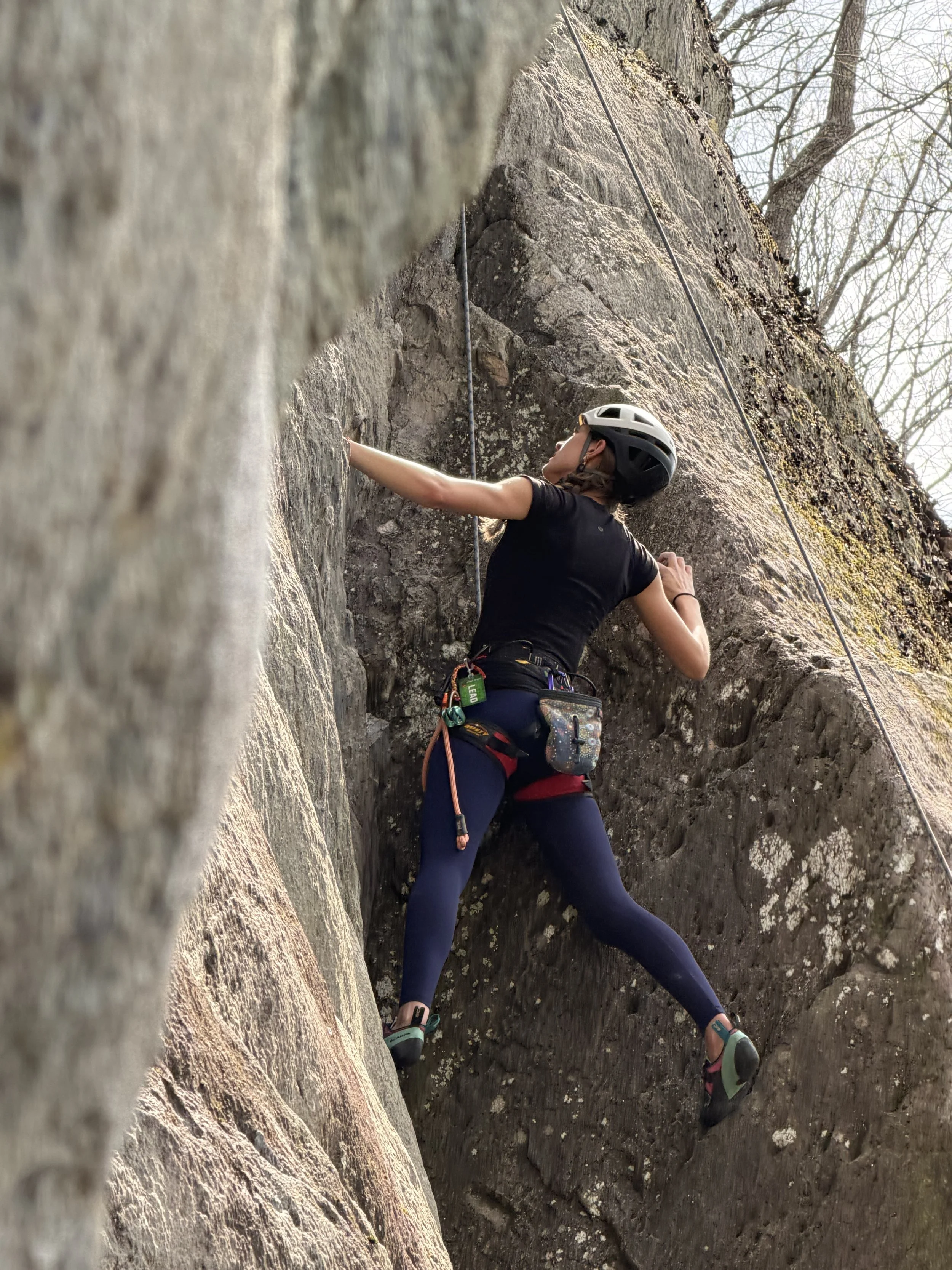 Two rock climbers wearing helmets sitting and standing on a rocky cliff, with a scenic view of autumn foliage and mountains in the background.