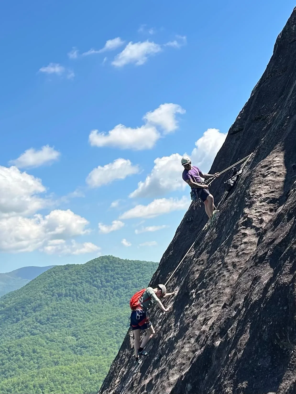 Two climbers ascending a steep rock face with safety gear, harnesses, and ropes, set against a backdrop of blue sky, clouds, and green mountains.