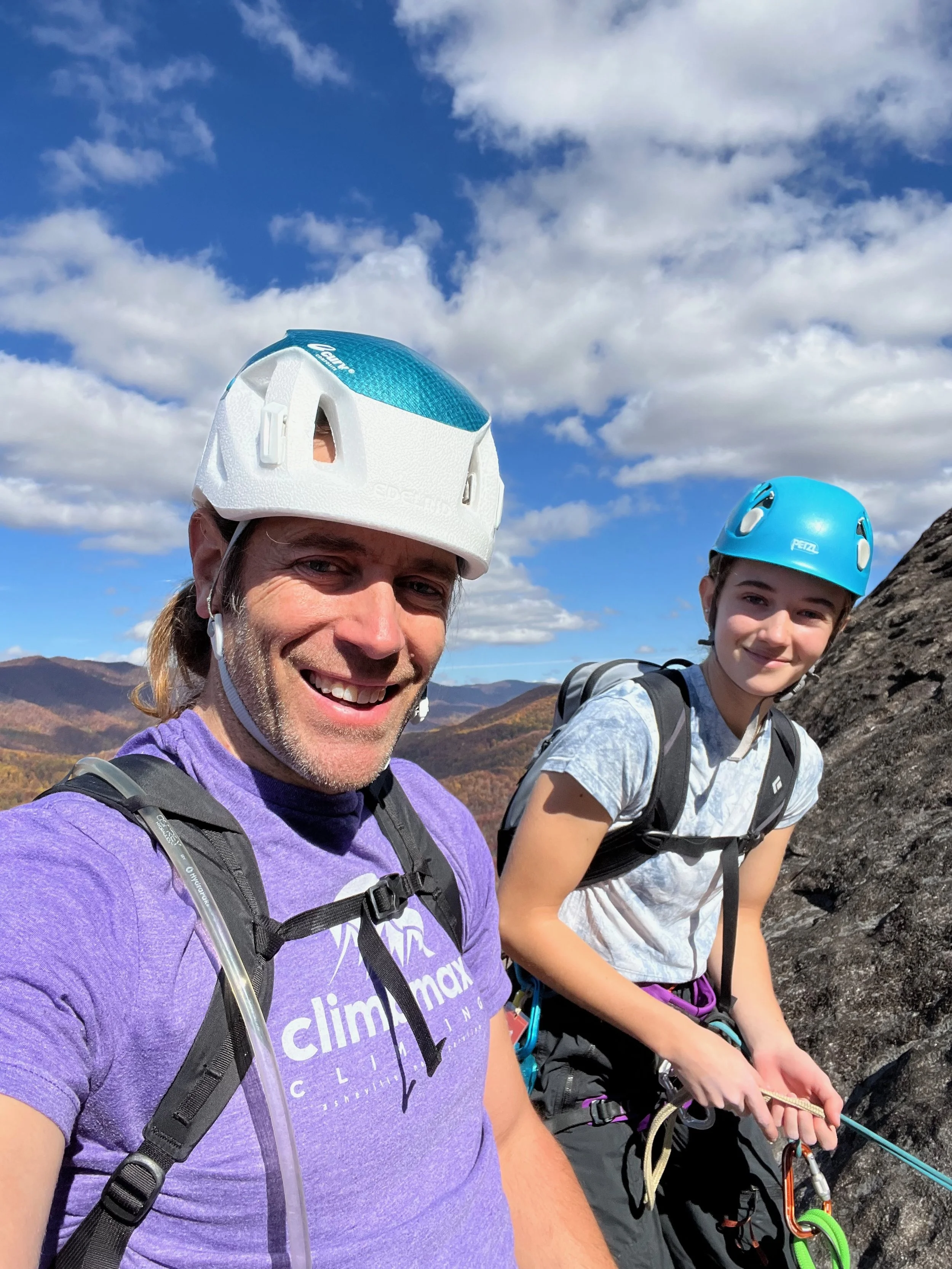 Two people rock climbing outdoors against a mountain landscape with a bright blue sky and clouds. They are wearing helmets and backpacks, and one is holding a climbing rope.