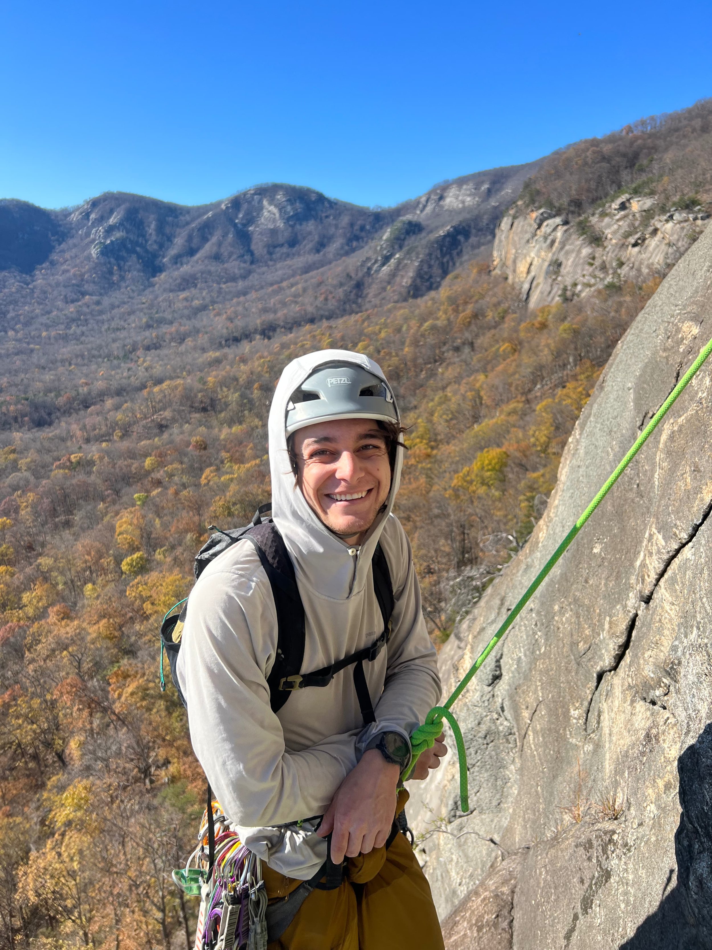 A man with a helmet climbing a rock face at Rumbling Bald outdoors with a background of mountains and trees in autumn colors.