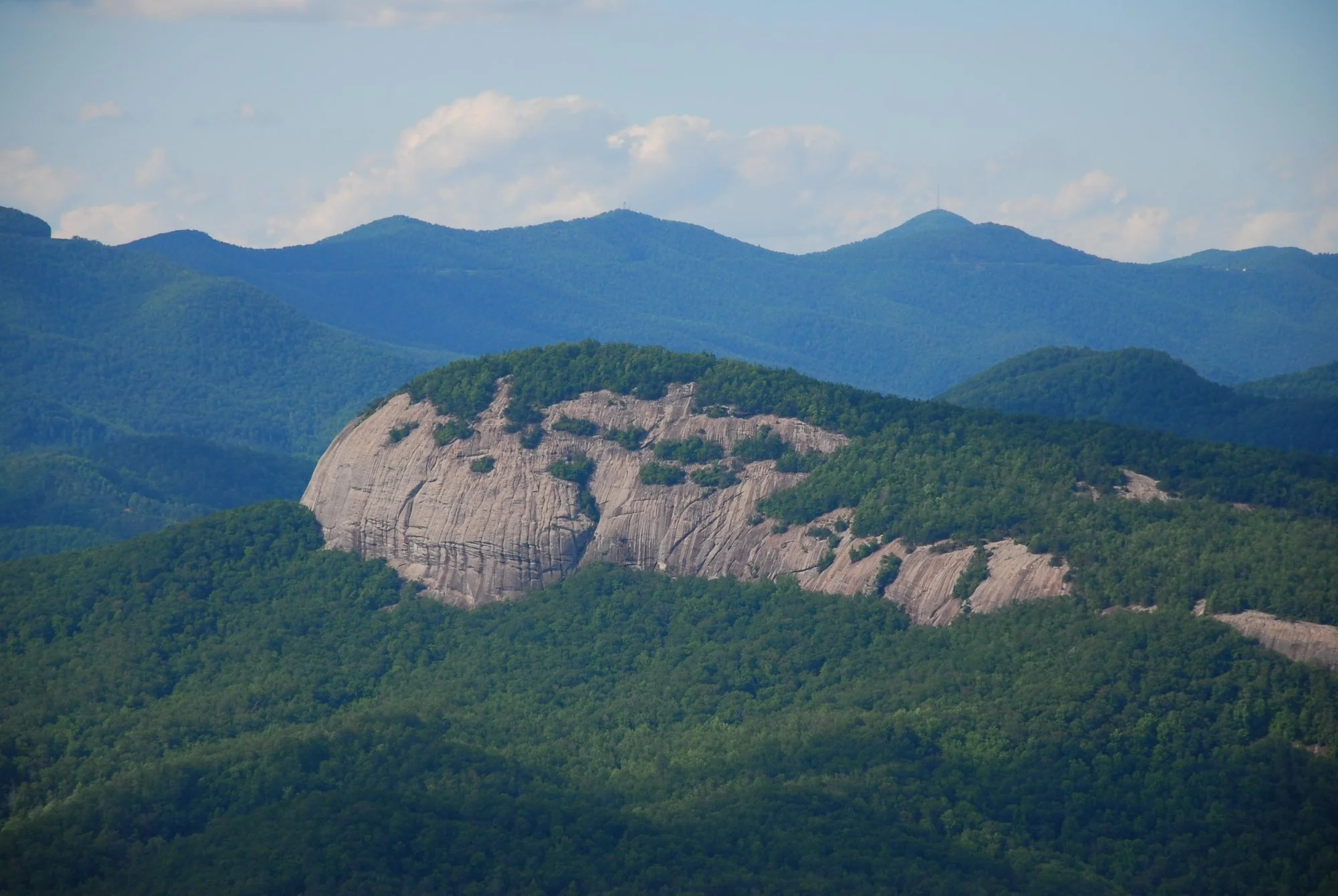View of a large rock formation with green forested hills and distant blue mountains under a partly cloudy sky.