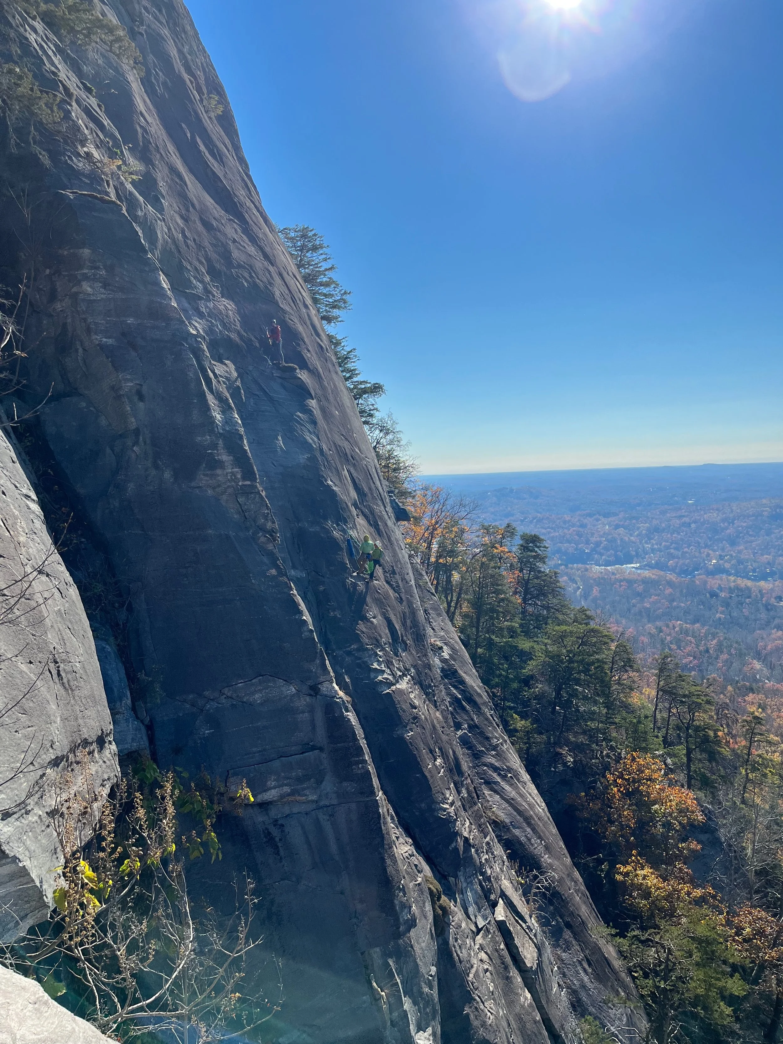 A rock climber ascending a steep granite cliff on Rumbling Bald, with a vast landscape of trees and a clear blue sky in the background.