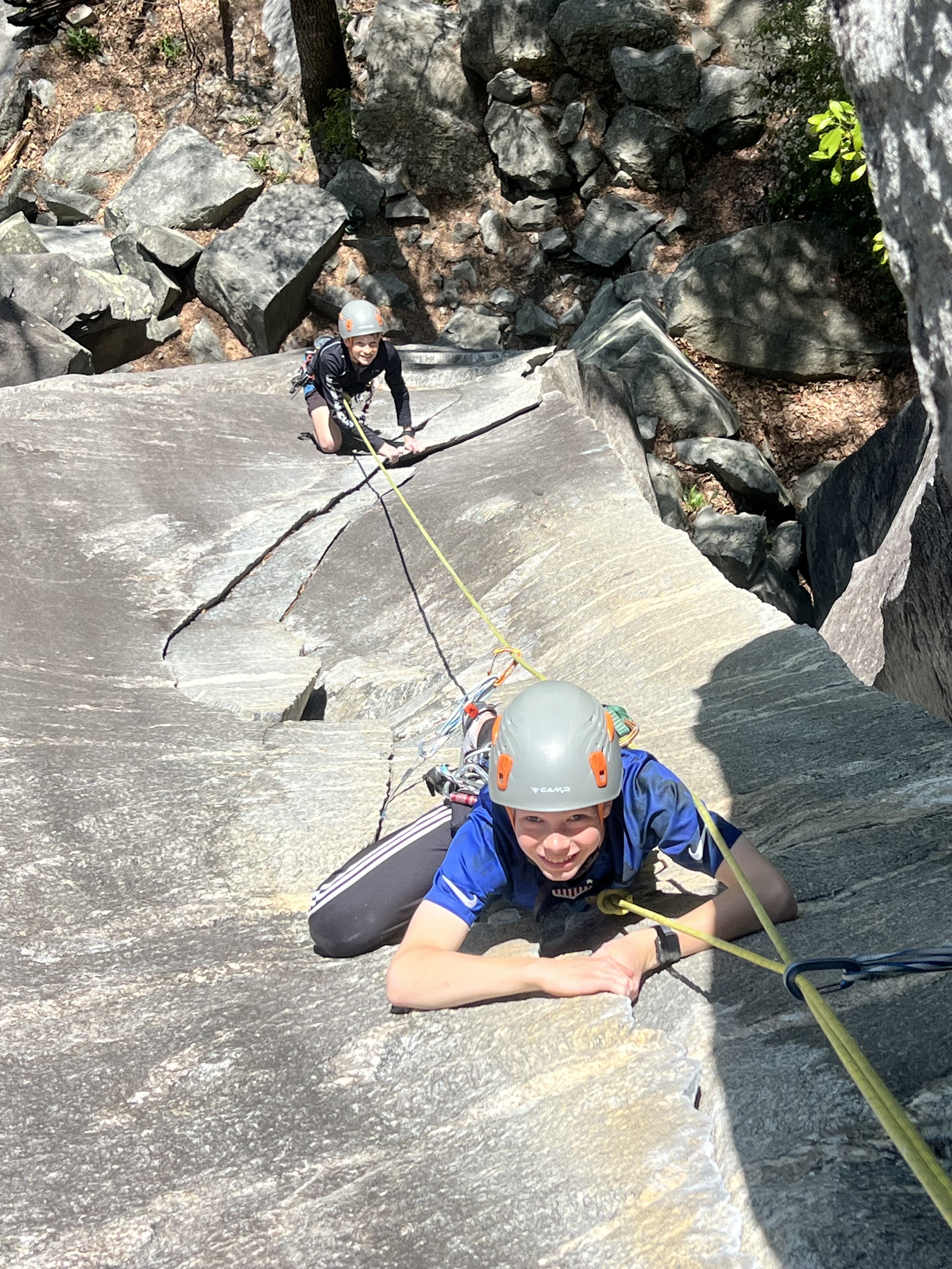 A young person rock climbing at Rumbling Bald, smiling, in a rocky outdoor setting, wearing a helmet and harness, taken from their perspective.