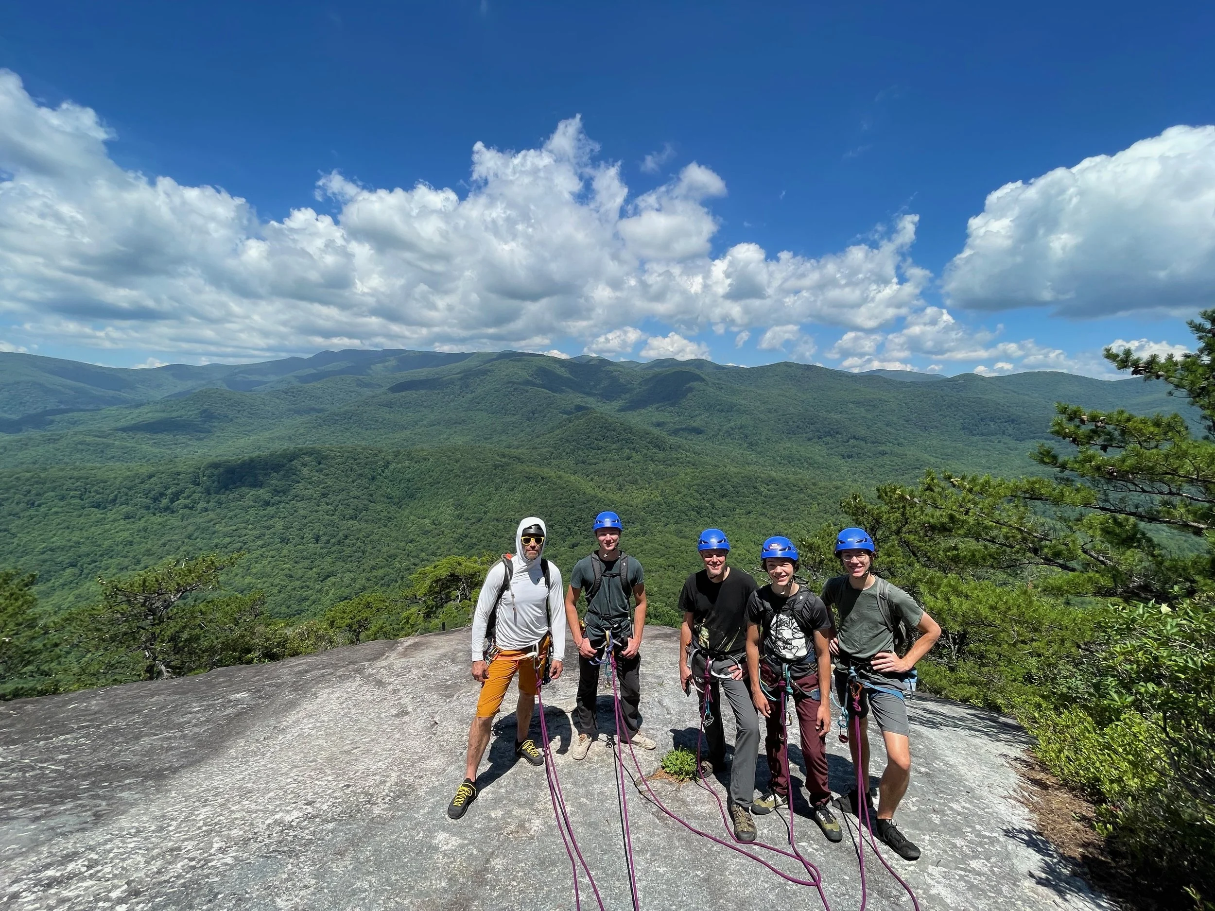Group of five people wearing helmets and harnesses standing on a rocky terrain with a lush green mountain range and blue sky with clouds in the background.