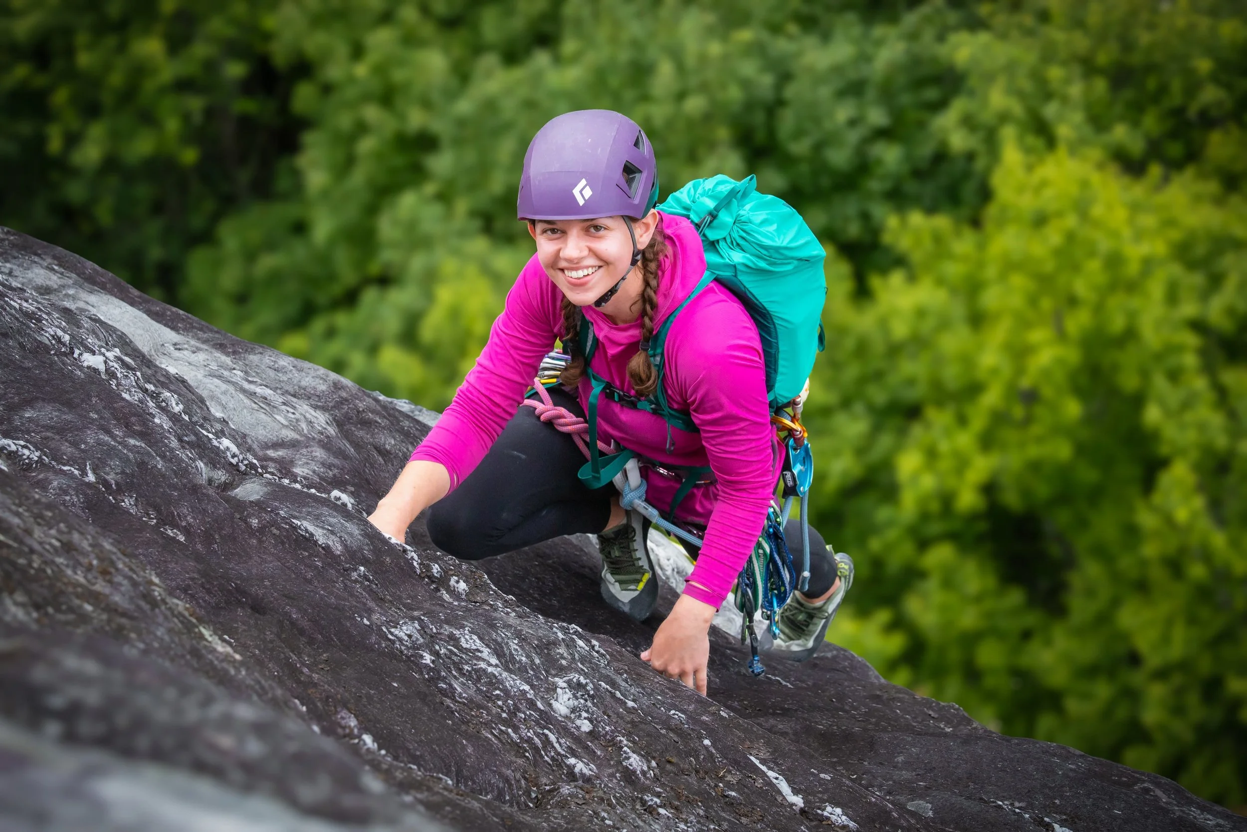 Young woman in pink jacket and purple helmet climbing a rock face outdoors, with a green forest in the background.