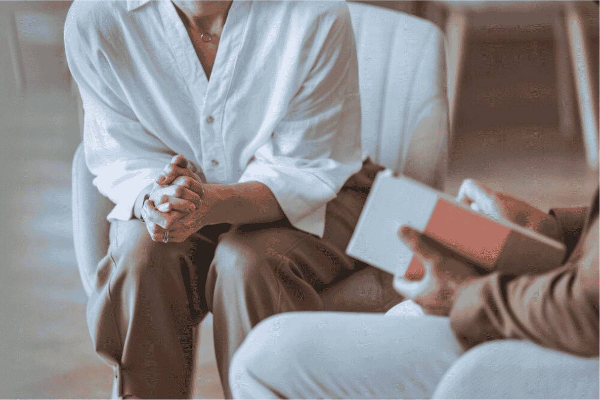 A person in a white shirt sitting with hands clasped, talking to another person holding a notebook in a therapy or counseling session.