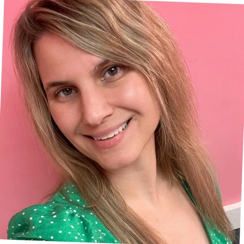 A woman with blonde hair wavy hair smiling, wearing a green polka-dot shirt, against a pink background.