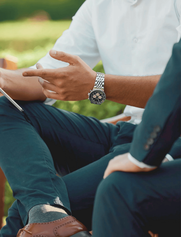 Close-up of two people sitting outdoors, one pointing and wearing a silver watch with a black dial, both dressed in formal attire.